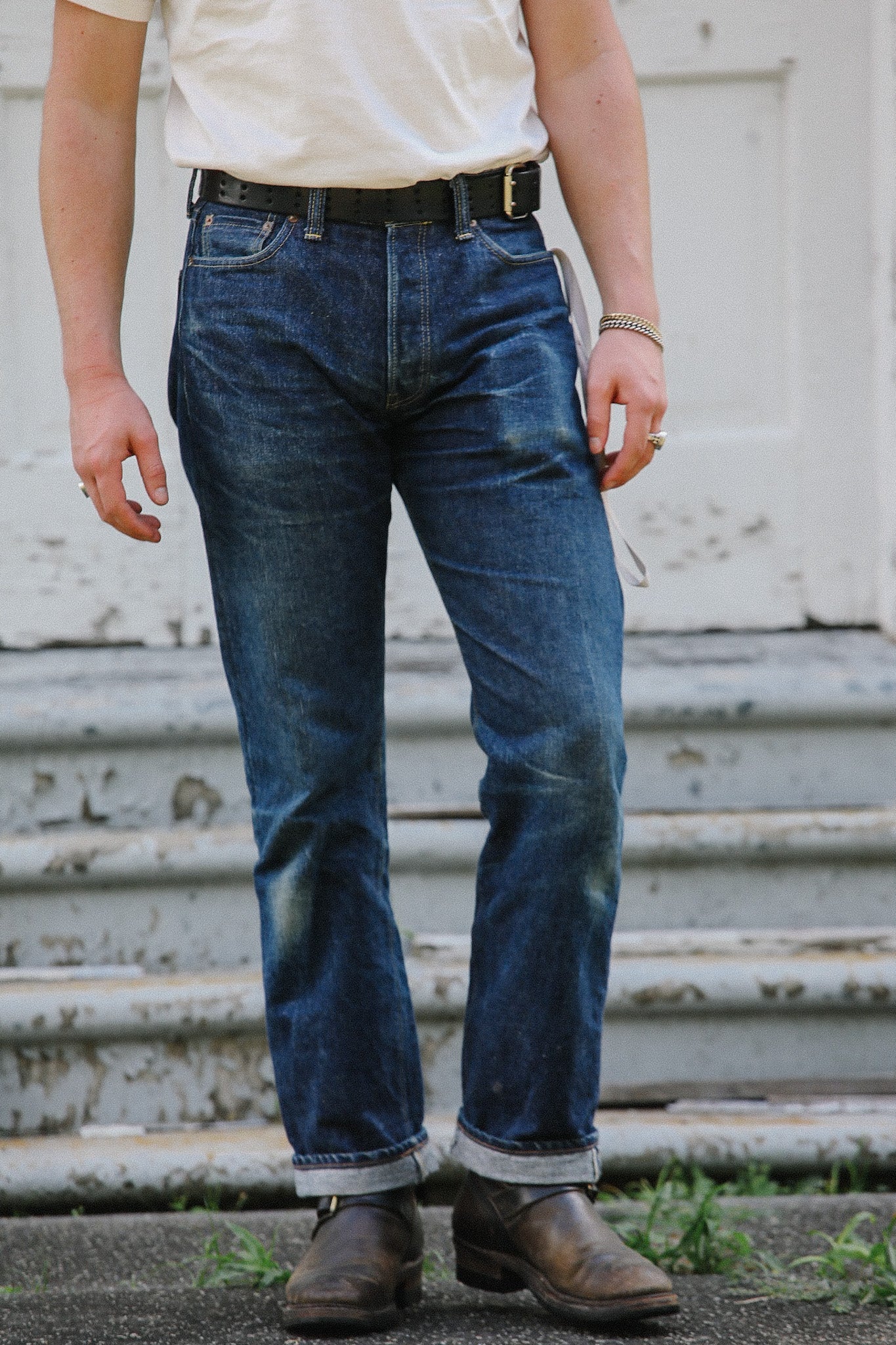 Person wearing rolled-up dark blue denim jeans with brown leather boots, white t-shirt, and black belt outdoors