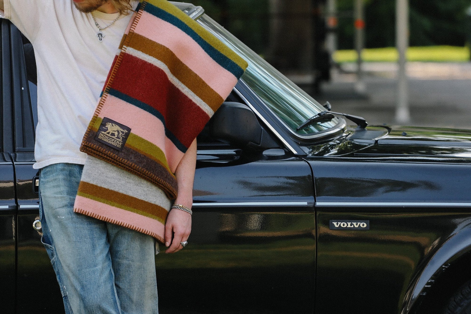 Person wearing a white shirt and jeans holding a striped Indigofera wool blanket leaning against a black Volvo car