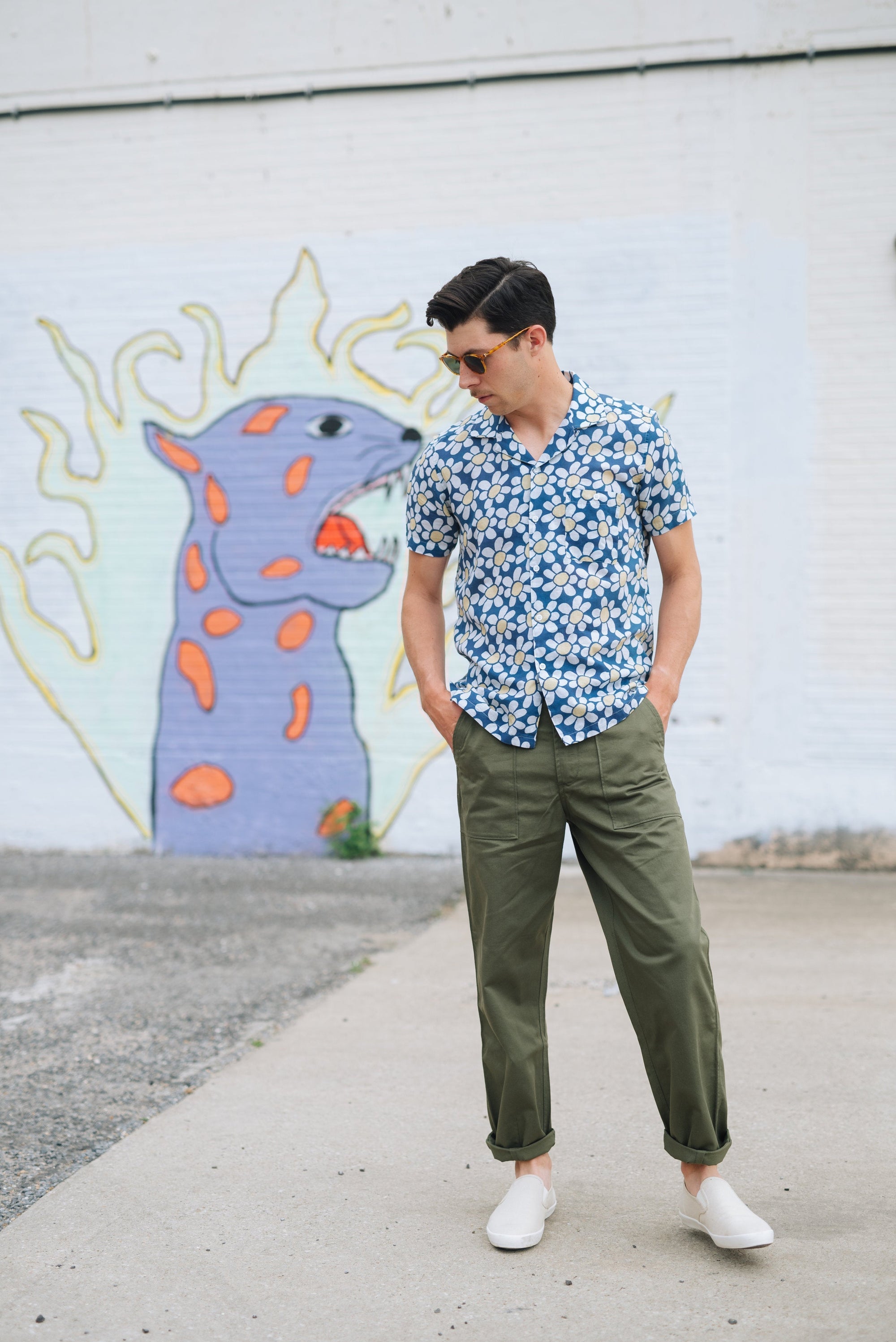 Man wearing floral print short sleeve shirt and olive green pants standing near colorful mural on white brick wall