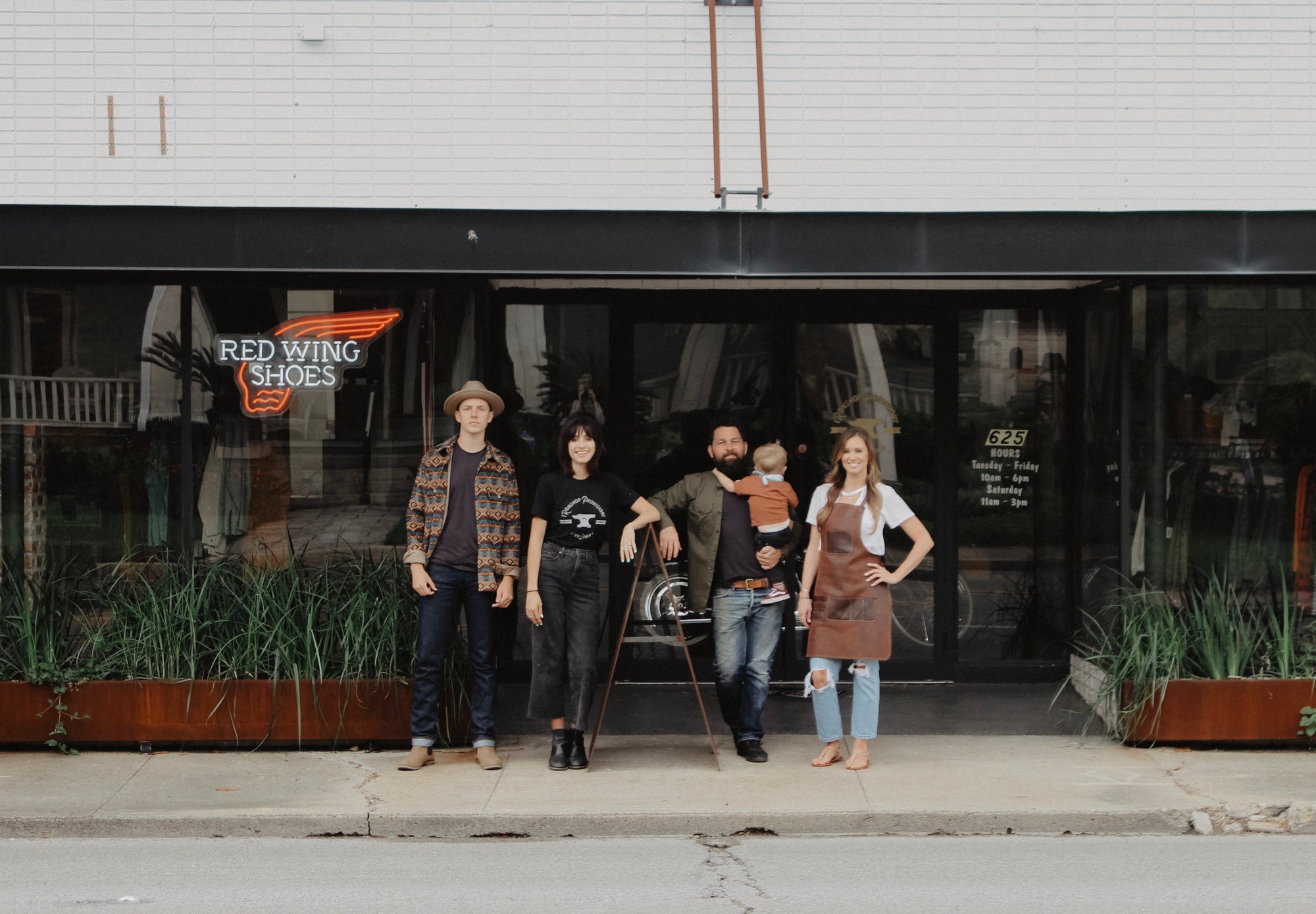 Four people standing outside Iron Shop Provisions store with Red Wing Shoes neon sign and storefront plants