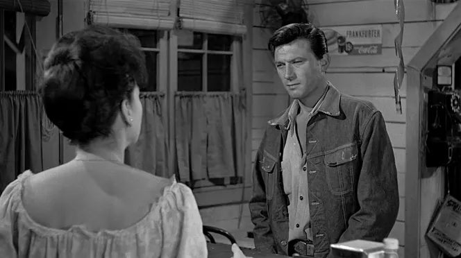 Black and white scene of a man in denim jacket talking to a woman in a rustic room with wooden walls