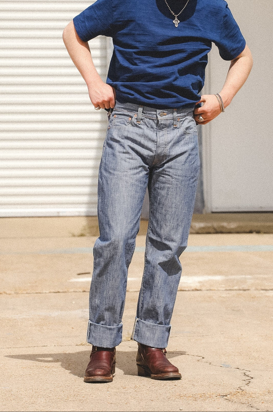 A man wearing a blue crewneck t-shirt, light-wash blue jeans, brown leather boots, and a gray baseball cap with a red logo, standing in front of a white garage door.