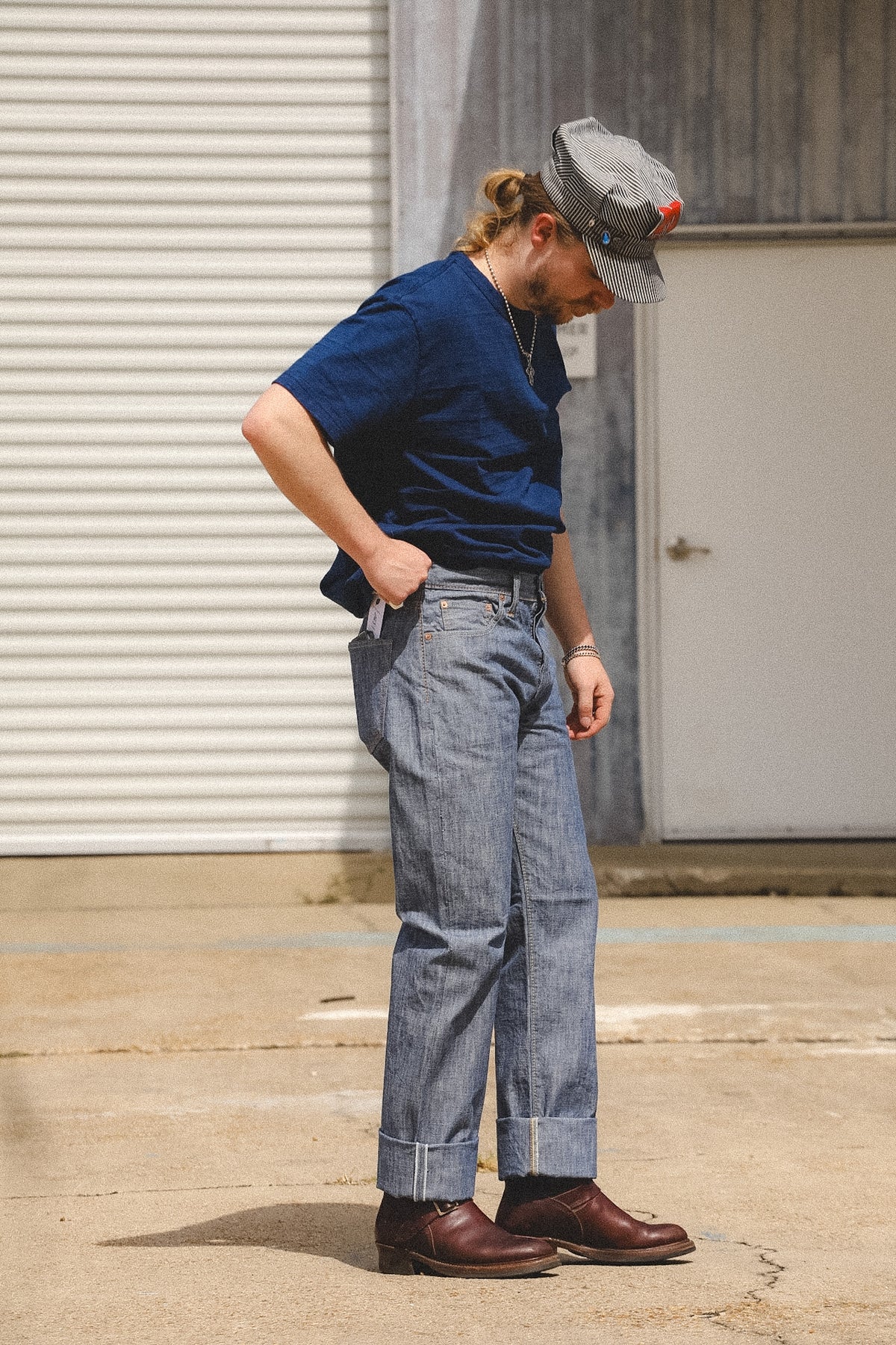 A man wearing a navy blue t-shirt, light-wash blue jeans, brown leather shoes, and a gray cap stands outdoors in front of a white garage door.