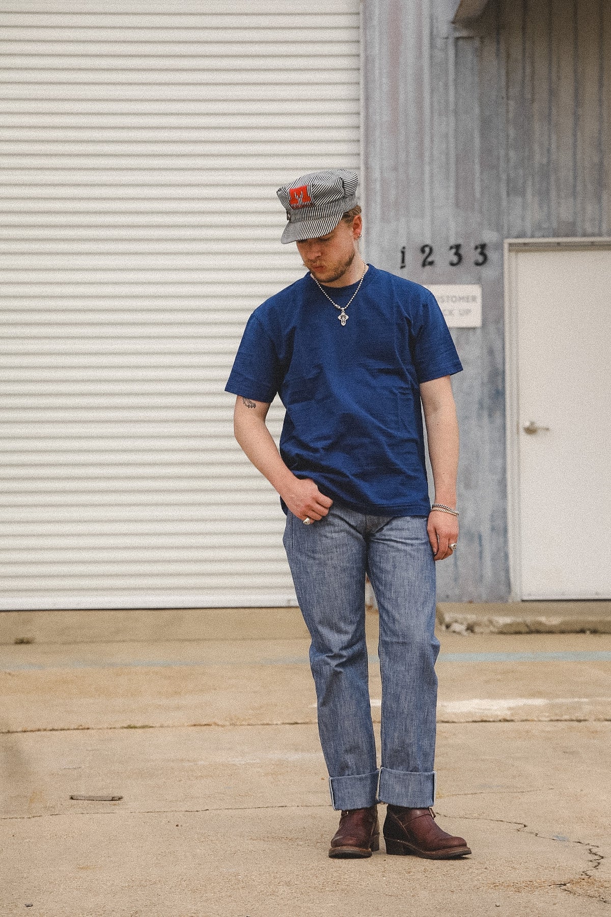 A man wearing a navy blue crewneck t-shirt, blue jeans, brown boots, and a gray baseball cap with a red logo, standing in front of a gray metal building.