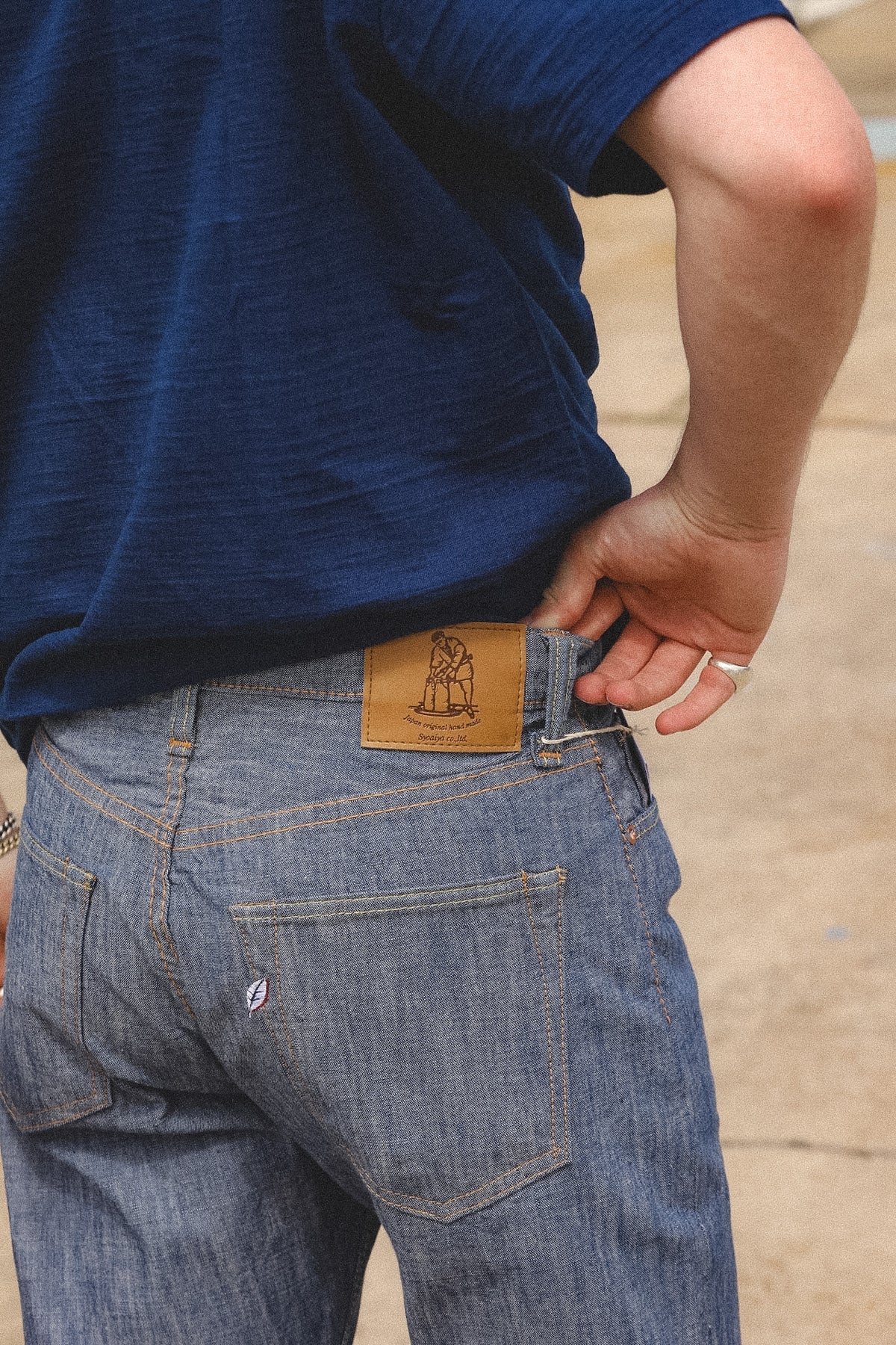 Close-up back view of a person wearing blue denim jeans and a navy T-shirt, pulling the waistband of the jeans to show a tan leather brand patch with a dog illustration.