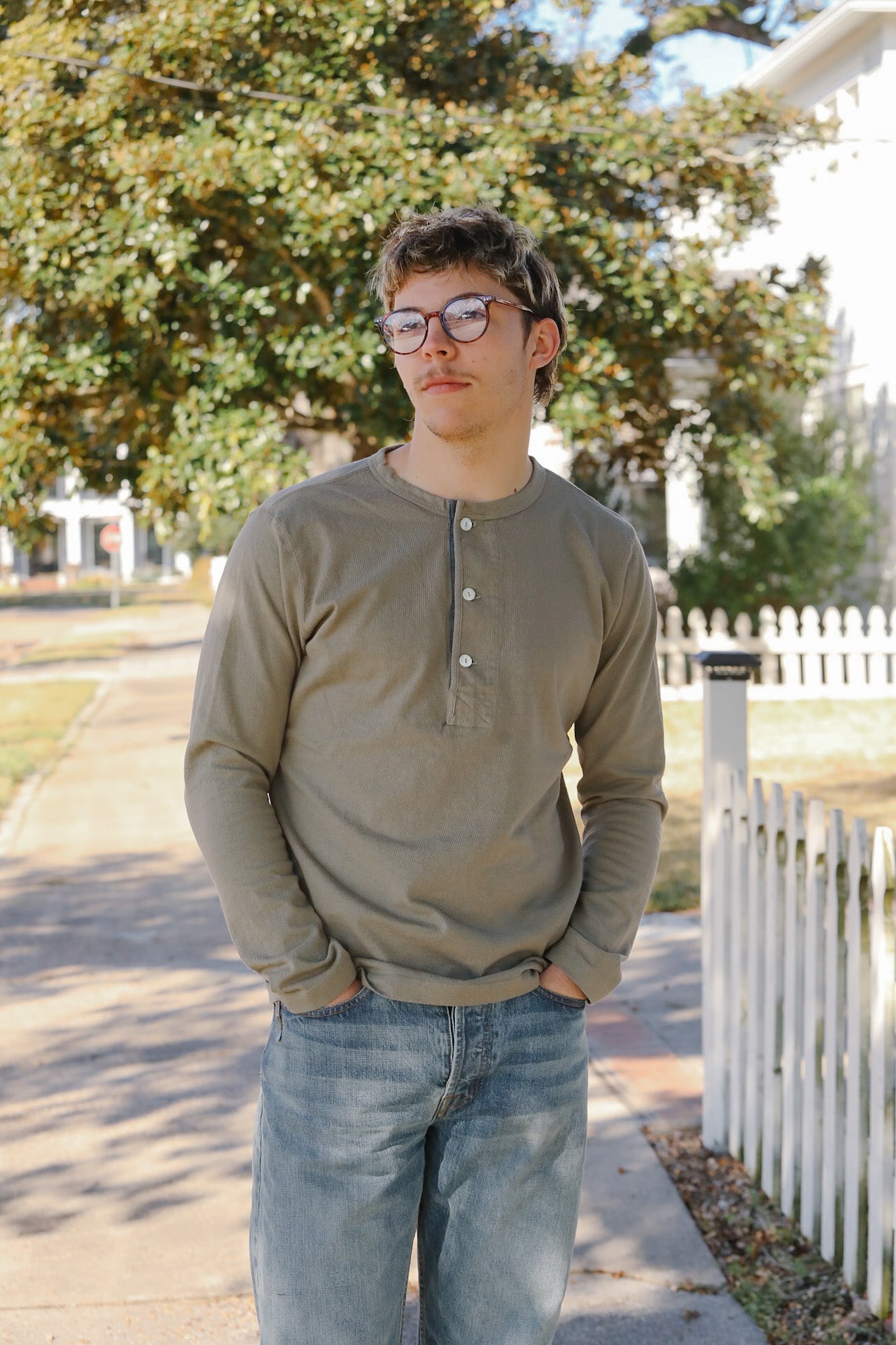 Man wearing glasses and a green shirt standing on a sidewalk with trees and a white fence in the background.