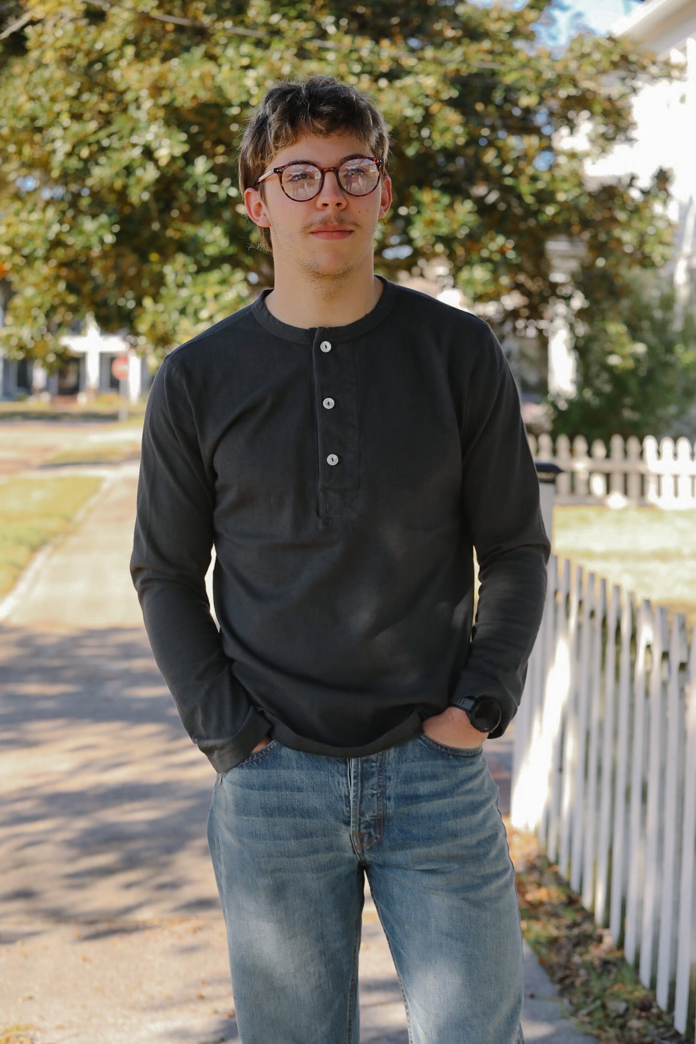 Person wearing a black long-sleeve shirt and blue jeans standing outdoors with trees and a white fence in the background.