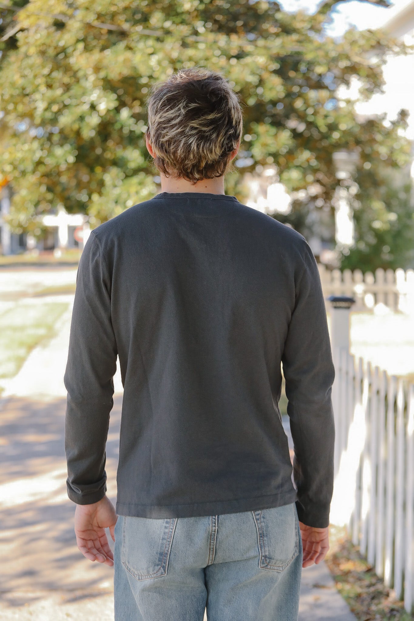 Man wearing a dark long-sleeve shirt and light blue jeans walking away from the camera with trees and a white fence in the background.