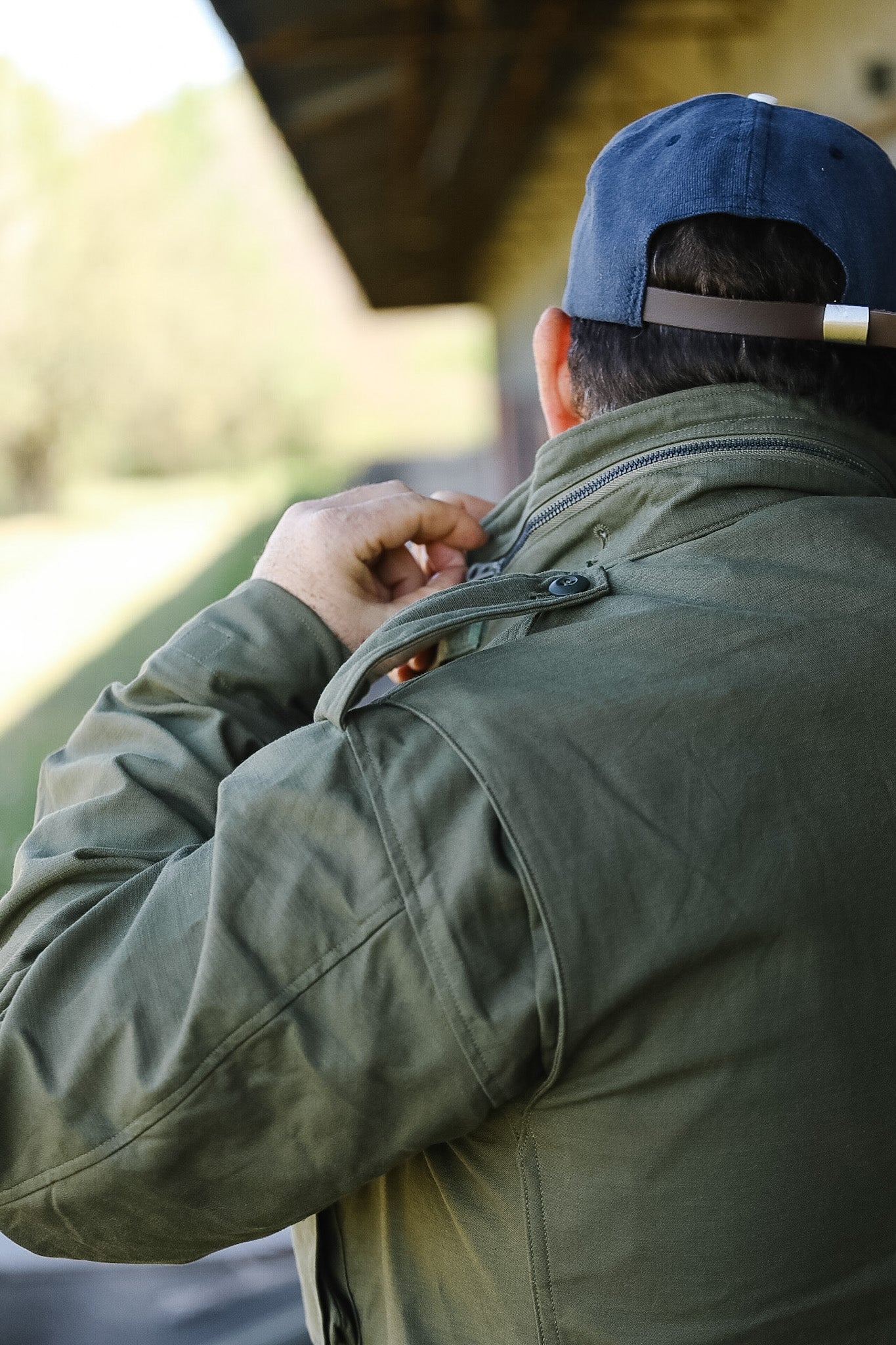Person wearing a green jacket and blue cap, adjusting something on their jacket outdoors.