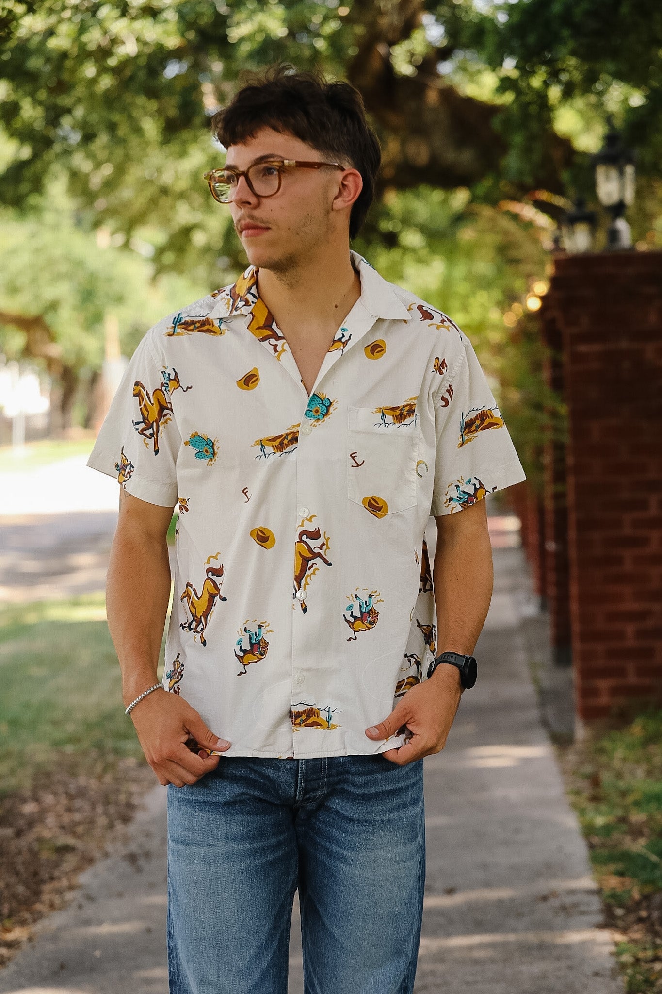 A man wearing a white short-sleeve button-up shirt with a colorful tropical and floral print, paired with blue jeans, standing outdoors on a sidewalk with trees and a brick wall in the background.