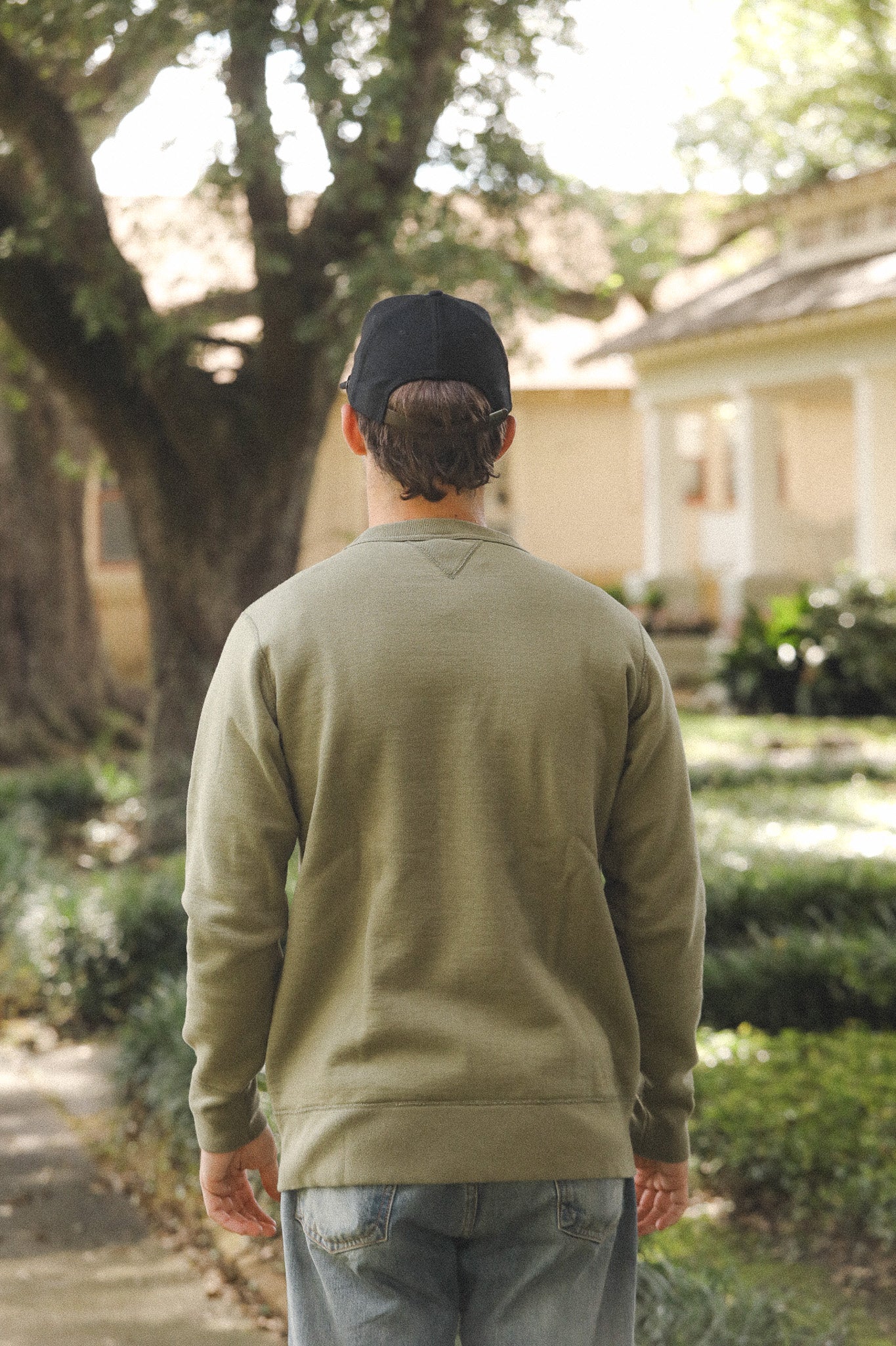 Person wearing a green sweatshirt and black cap walking away in a garden setting
