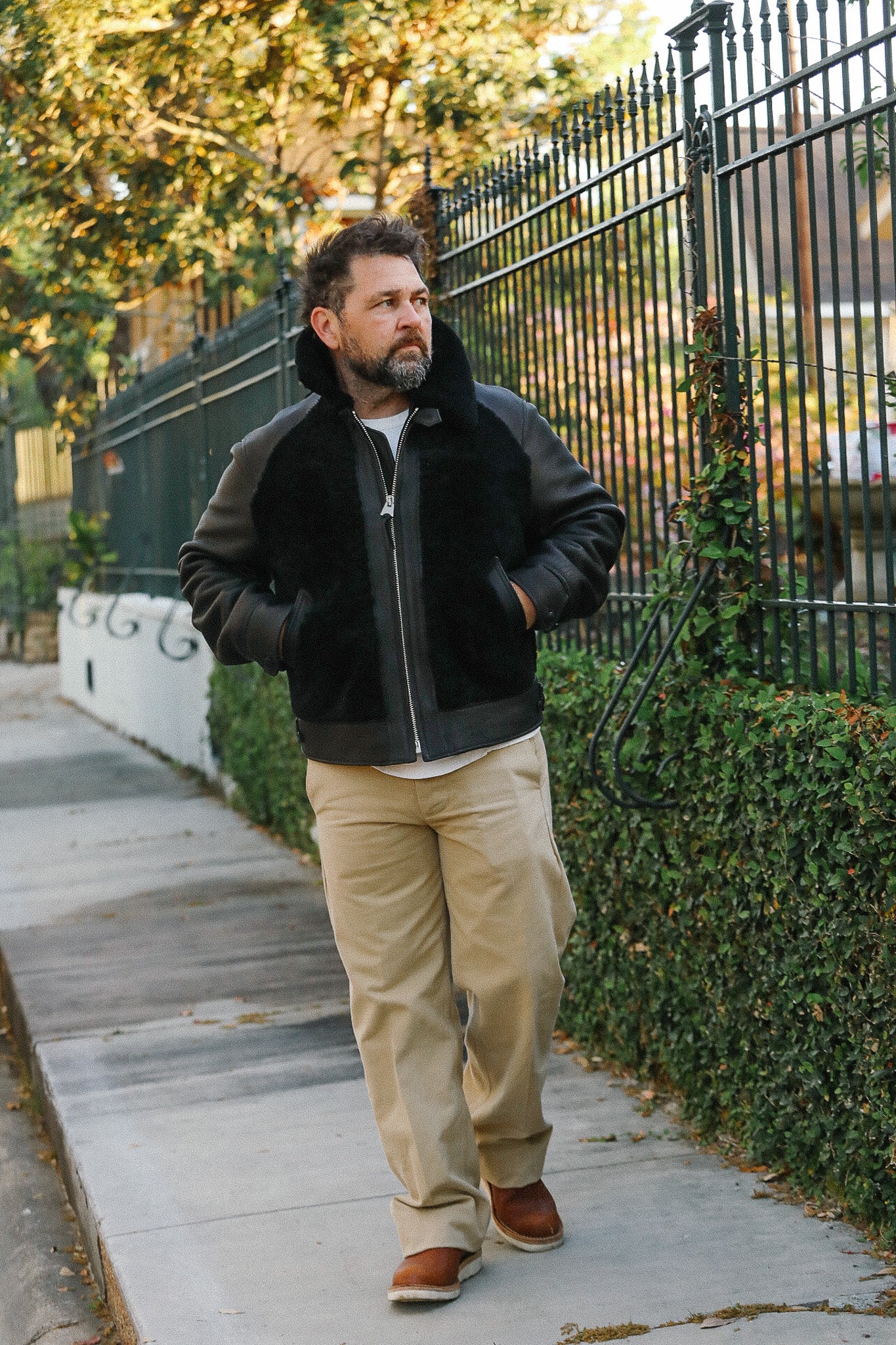 Man wearing a black jacket with fur lining, beige pants, and brown shoes walking on a sidewalk.