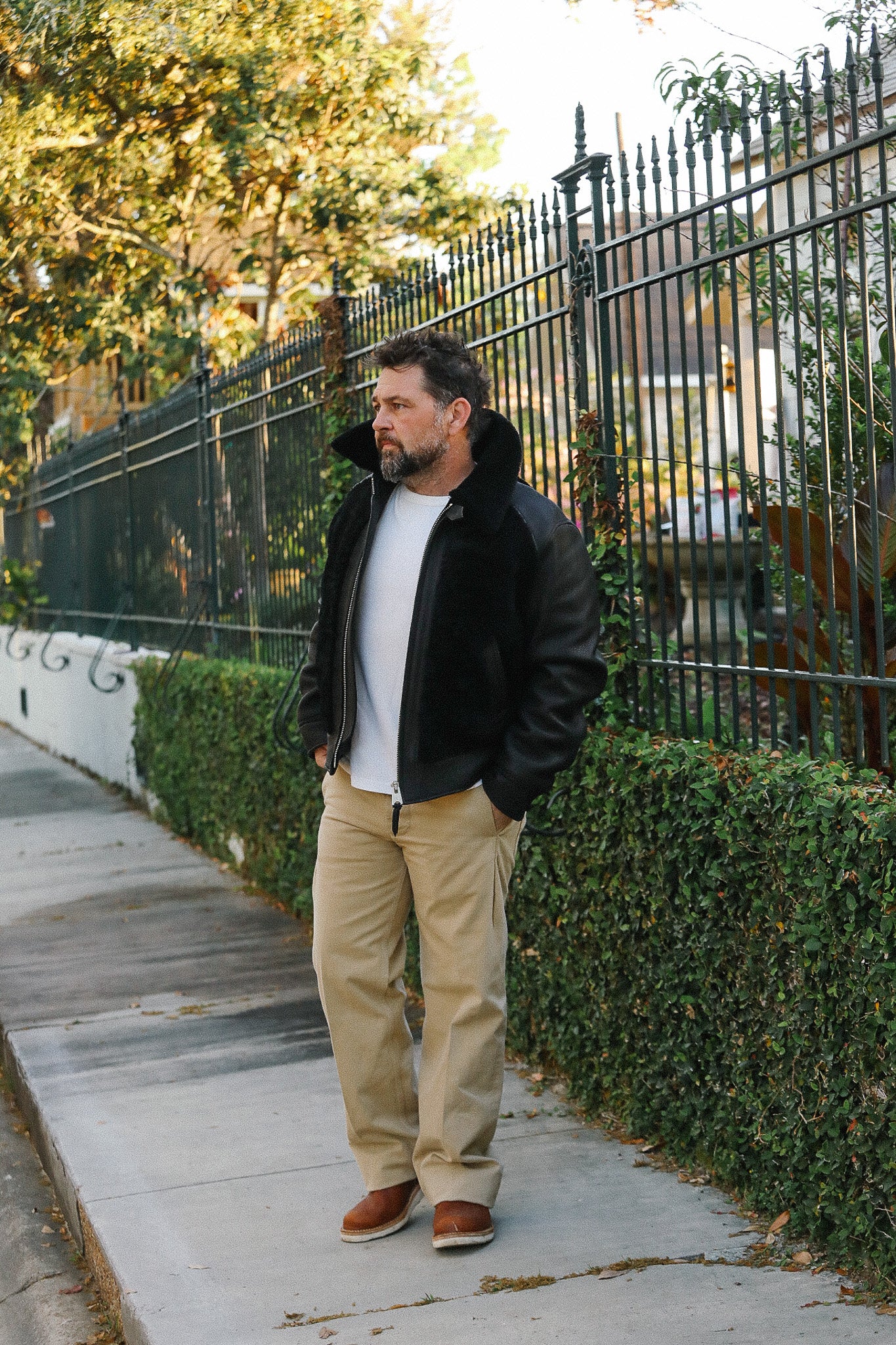 Man in black jacket and beige pants standing on a sidewalk with trees and a fence in the background