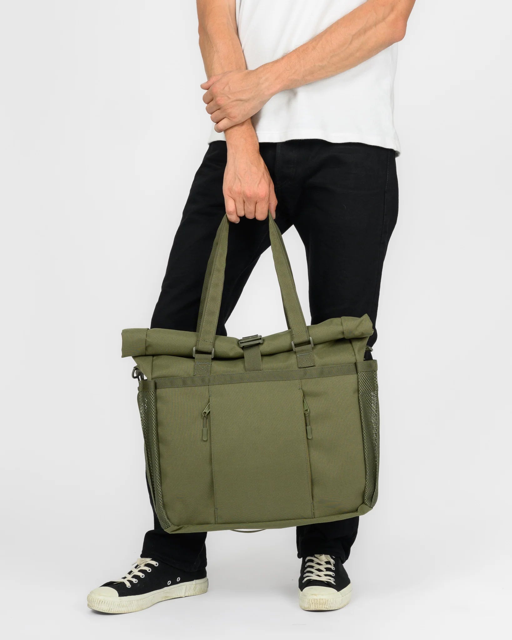 An olive green roll-top duffel bag with dual top handles, held by a model wearing a white t-shirt, black pants, and black sneakers against a light gray background.