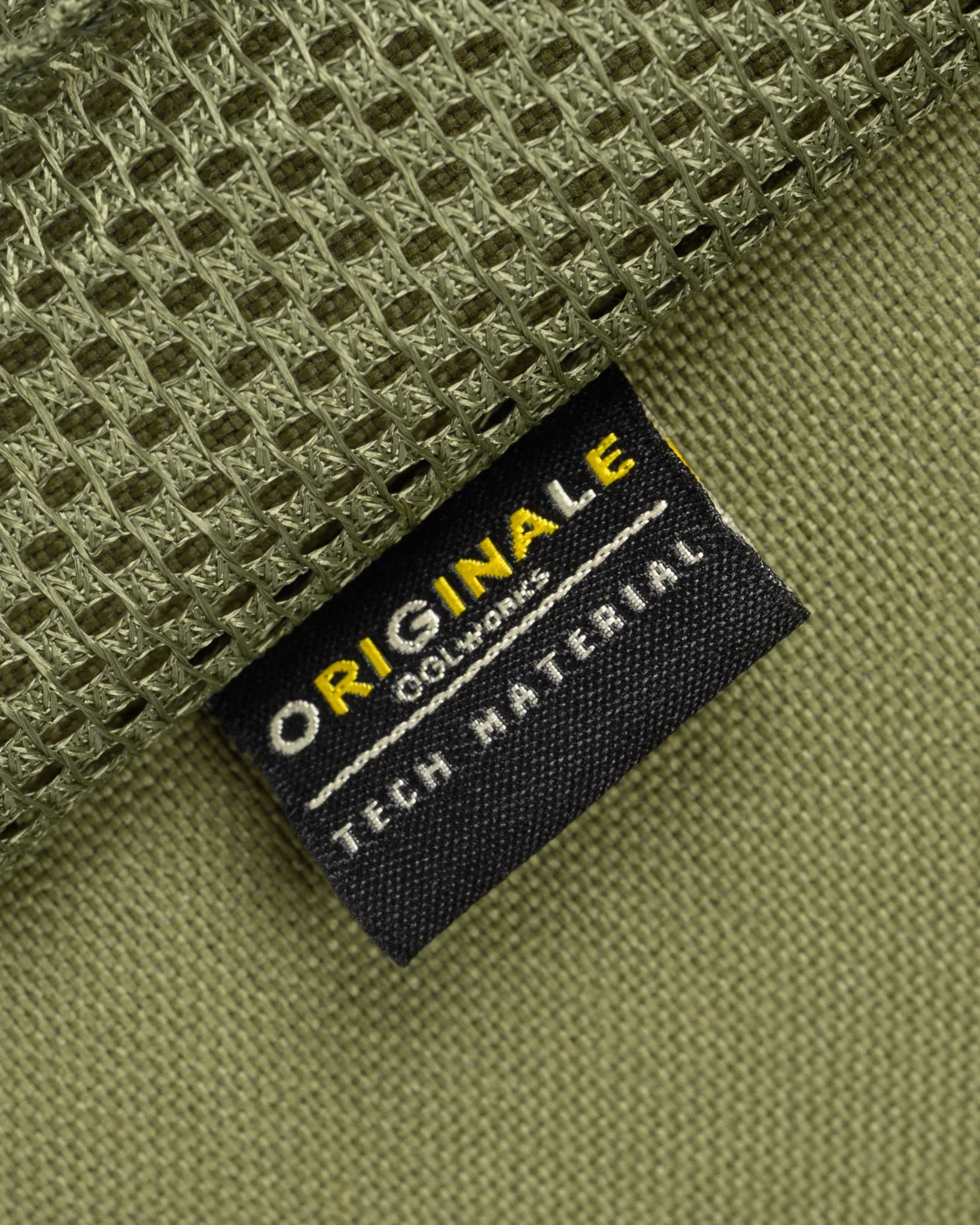 Close-up of a black fabric brand tag reading 'ORIGINE CO-WORKS TECH MATERIAL' in white and yellow text, attached to olive green technical mesh fabric.