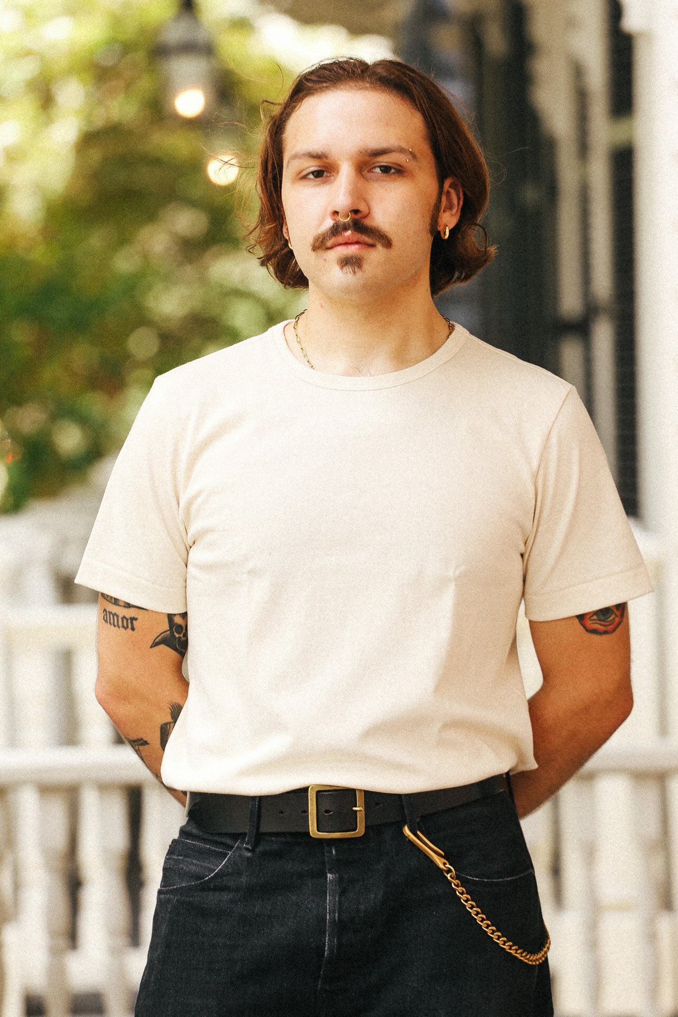 A man with a mustache and shoulder-length hair wearing a cream-colored crewneck t-shirt, dark jeans with a black belt, and a gold chain accessory, standing outdoors in front of a white railing and green foliage.