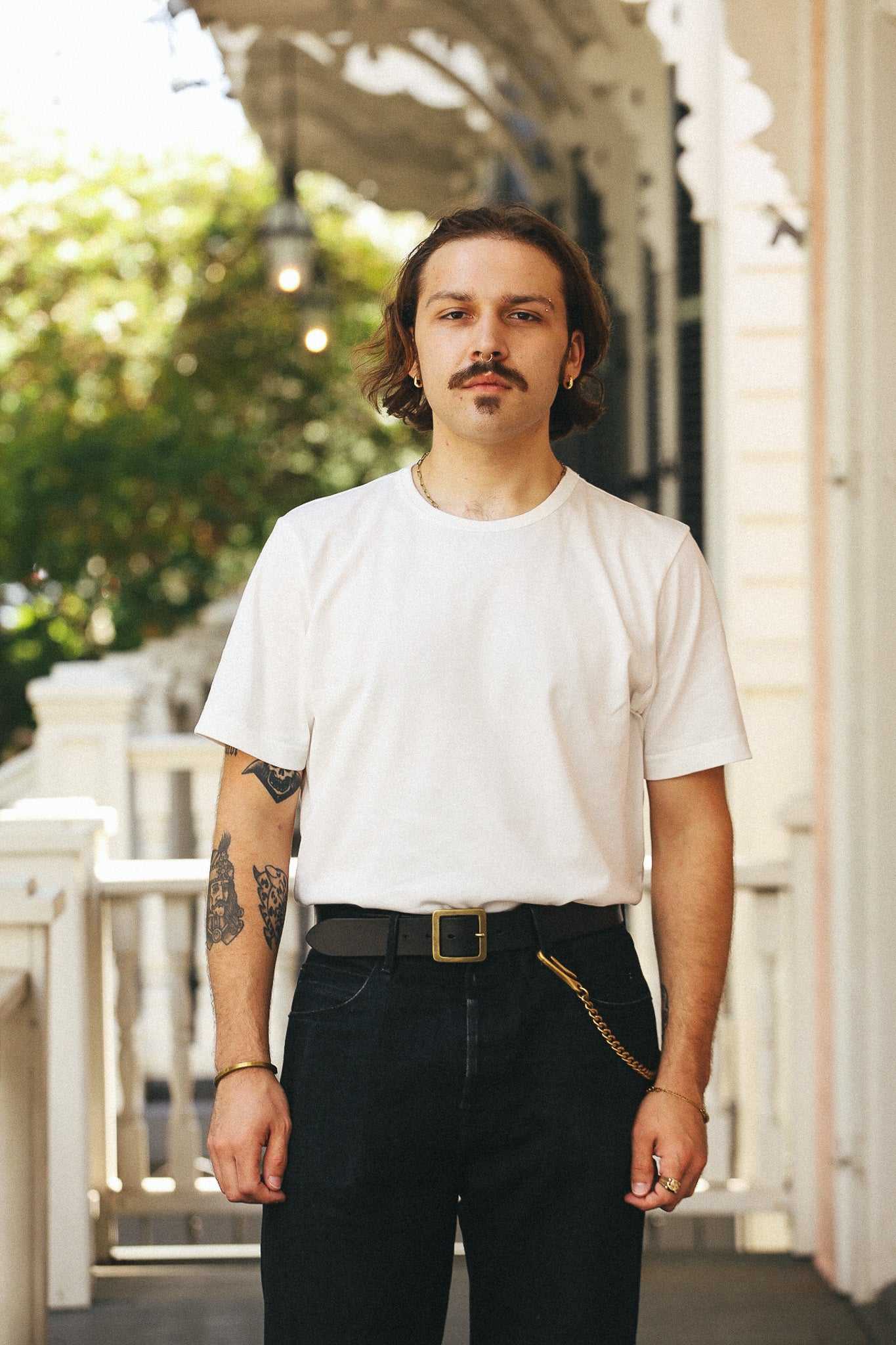 A man with a mustache and tattoos stands on a porch wearing a plain white crew-neck t-shirt, dark jeans, a black belt, and a gold chain accessory.