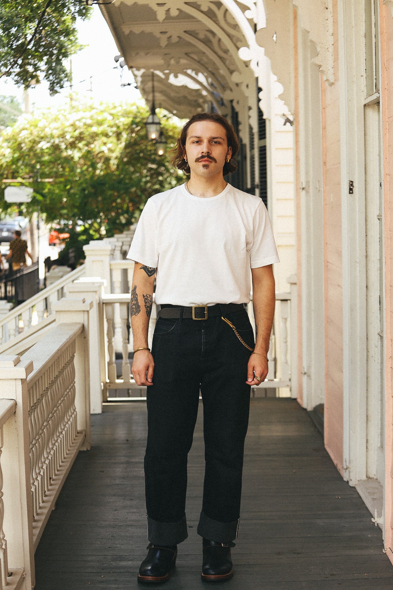 A man stands on a porch wearing a plain white crew-neck t-shirt, dark charcoal gray jeans with rolled cuffs, a black belt with a gold buckle, and black leather shoes.