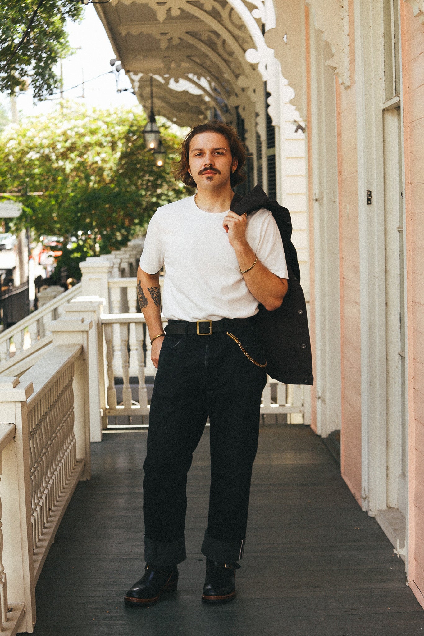 A man stands on a porch wearing a white crew-neck t-shirt, dark charcoal gray cuffed jeans, and black leather boots, with a black jacket slouched over his shoulder. The background features a white decorative porch railing and a pink building facade.