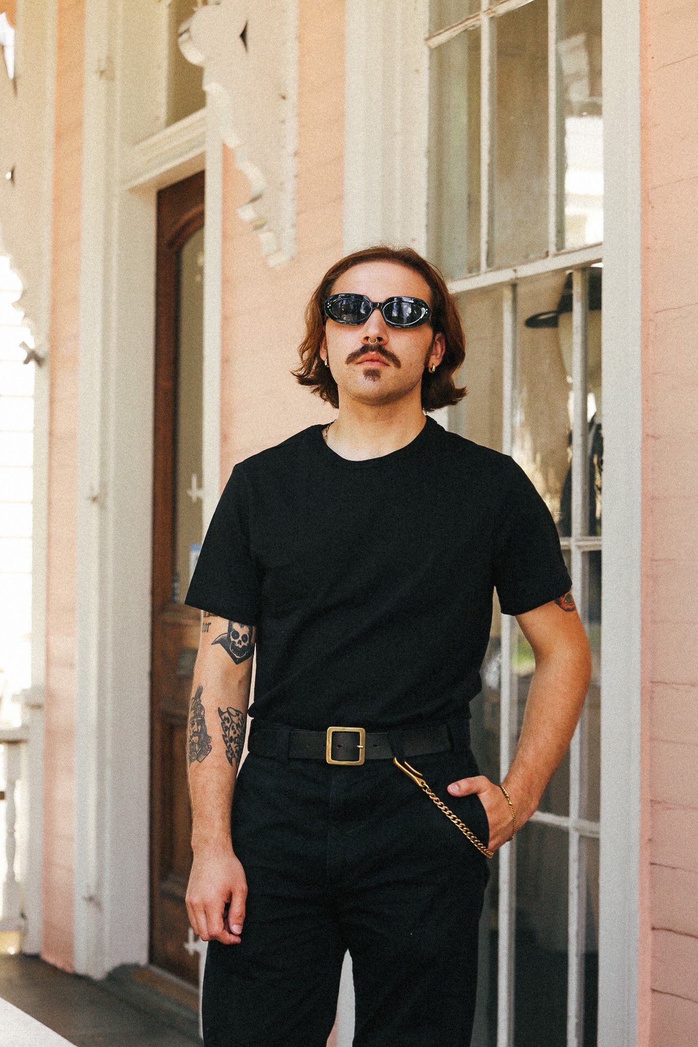 A man with a mustache and sunglasses wearing a black t-shirt, black pants, and a black belt with a gold buckle, standing in front of a pink building.