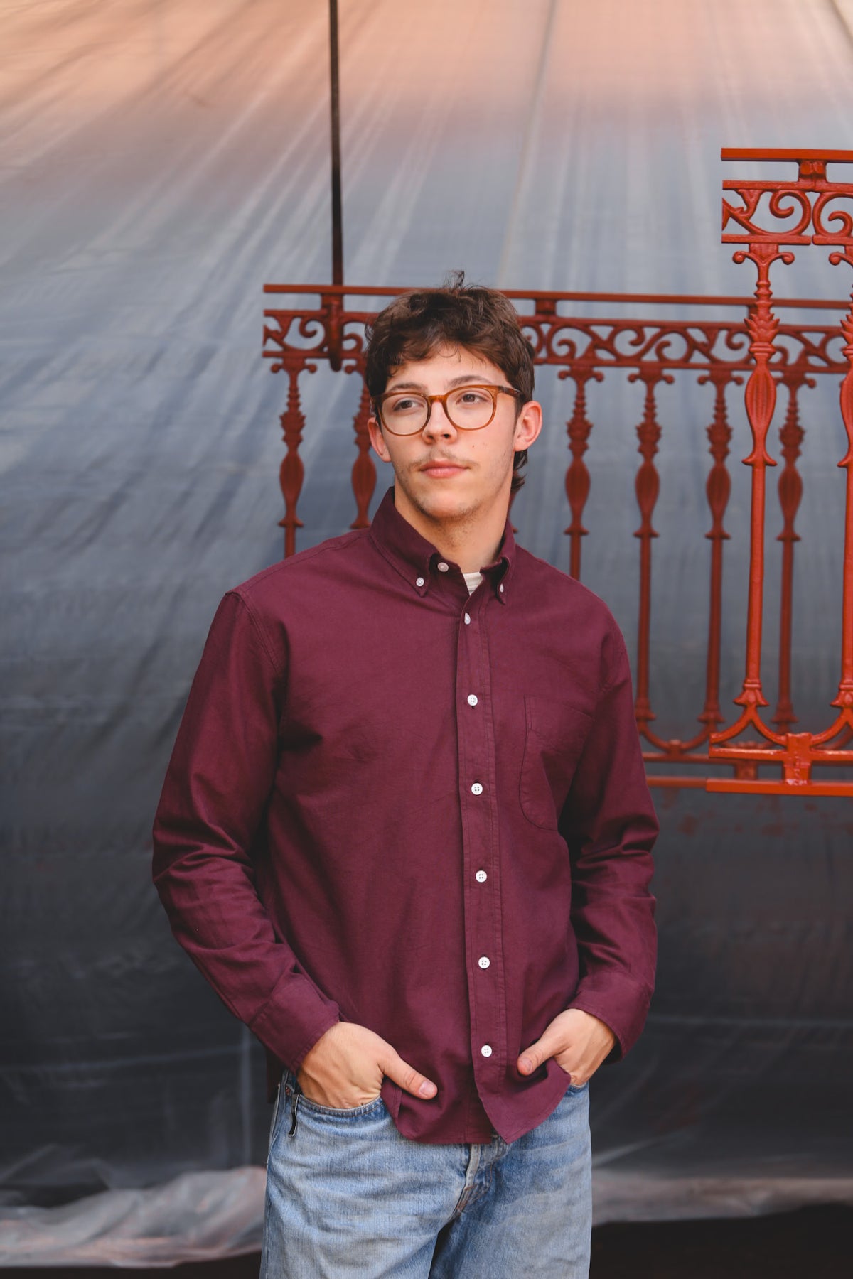 A young man with glasses wearing a maroon button-down shirt and blue jeans, standing in front of a gray fabric backdrop with a red metal railing.