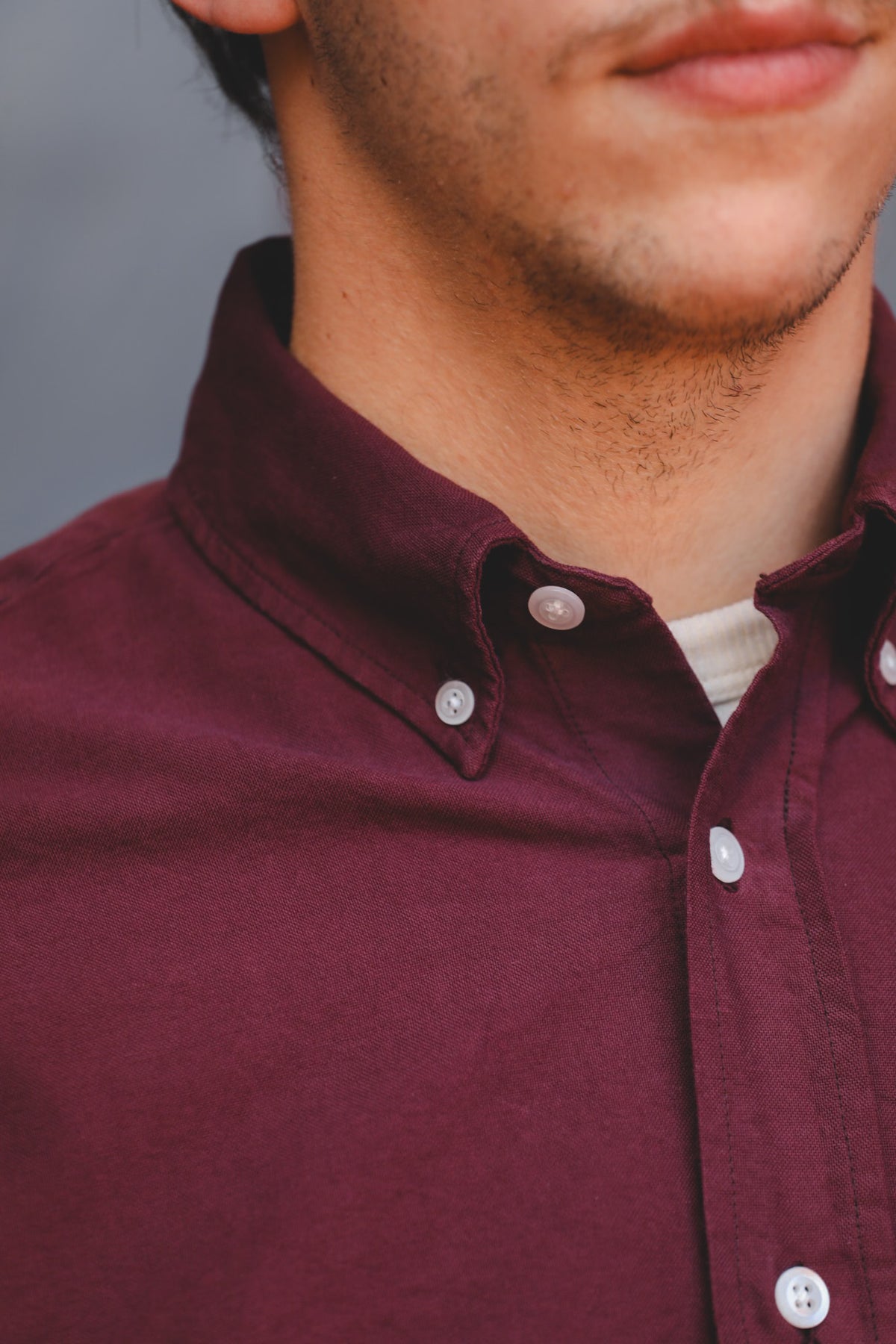 Close-up of a man wearing a burgundy button-down shirt with white buttons, showing the collar, chest area, and part of his chin and neck.
