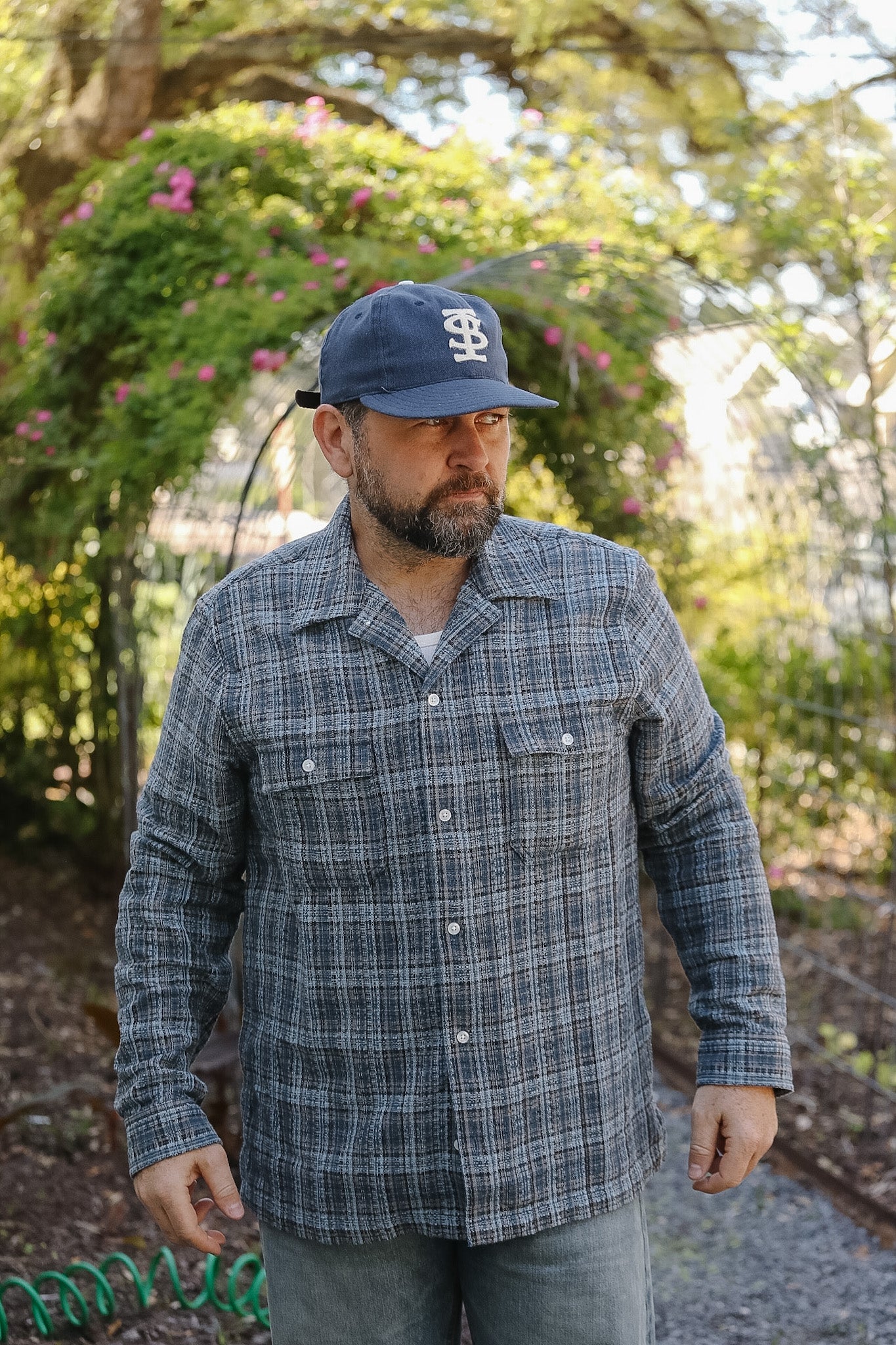 Man wearing a blue plaid shirt and cap outdoors with greenery in the background