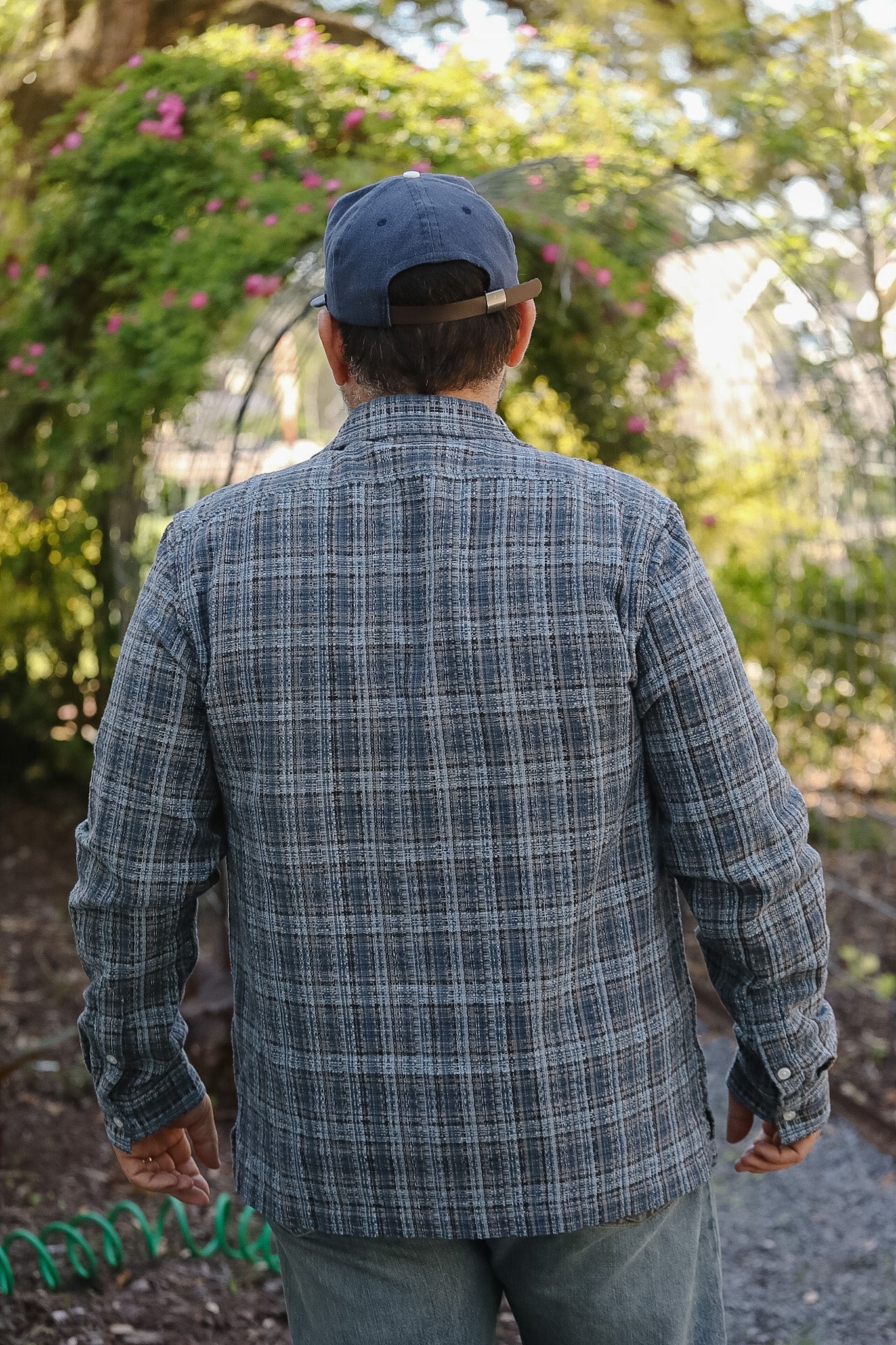 Man wearing a plaid shirt and cap, standing outdoors with greenery in the background