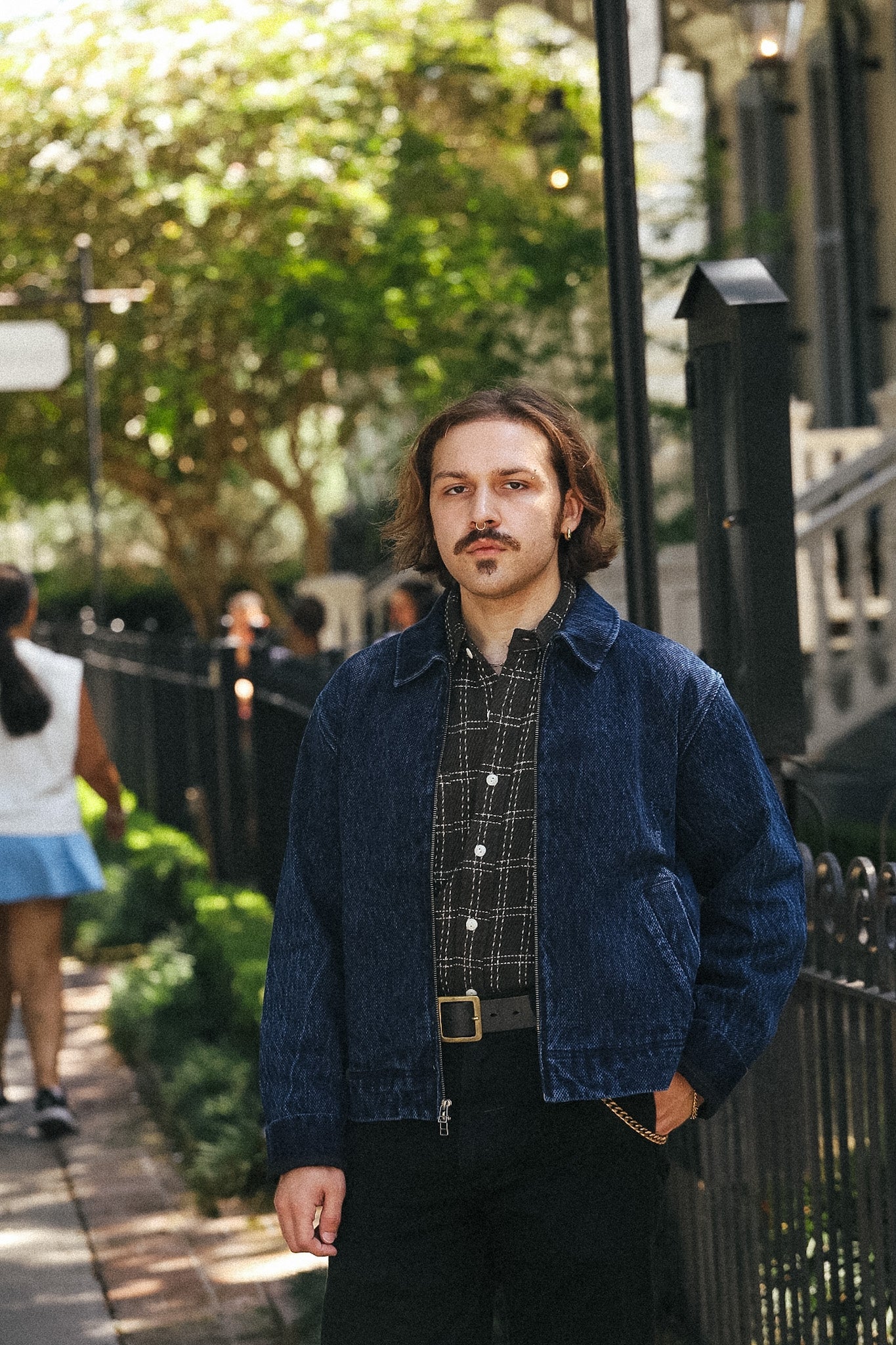 A man with a mustache stands on an outdoor city sidewalk wearing a dark blue corduroy jacket over a black and white patterned button-down shirt, paired with black pants and a gold-buckled belt.