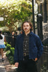 A man with a mustache stands on an outdoor city sidewalk wearing a dark blue corduroy jacket over a black and white patterned button-down shirt, paired with black pants and a gold-buckled belt.