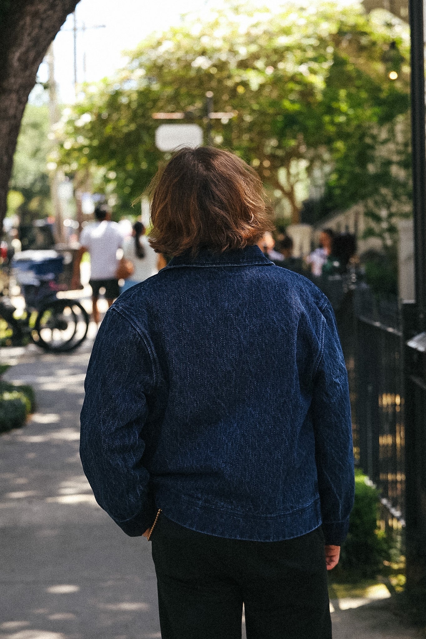 A person viewed from behind wearing a dark blue denim jacket and black pants, standing on a sunlit sidewalk with trees, pedestrians, and a fence in the background.
