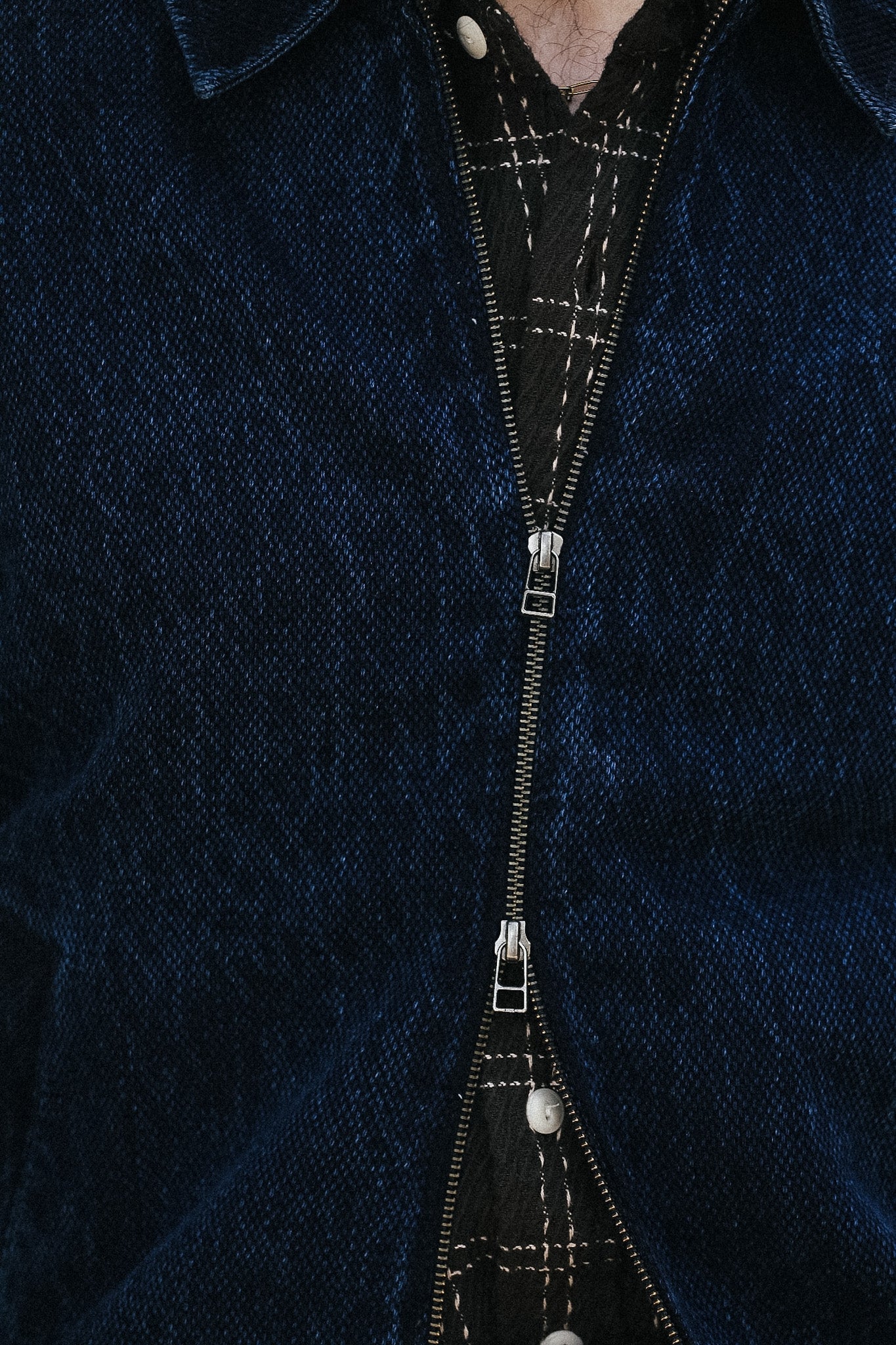 Close-up of a dark blue textured jacket or overshirt with a central silver zipper over a black plaid shirt.