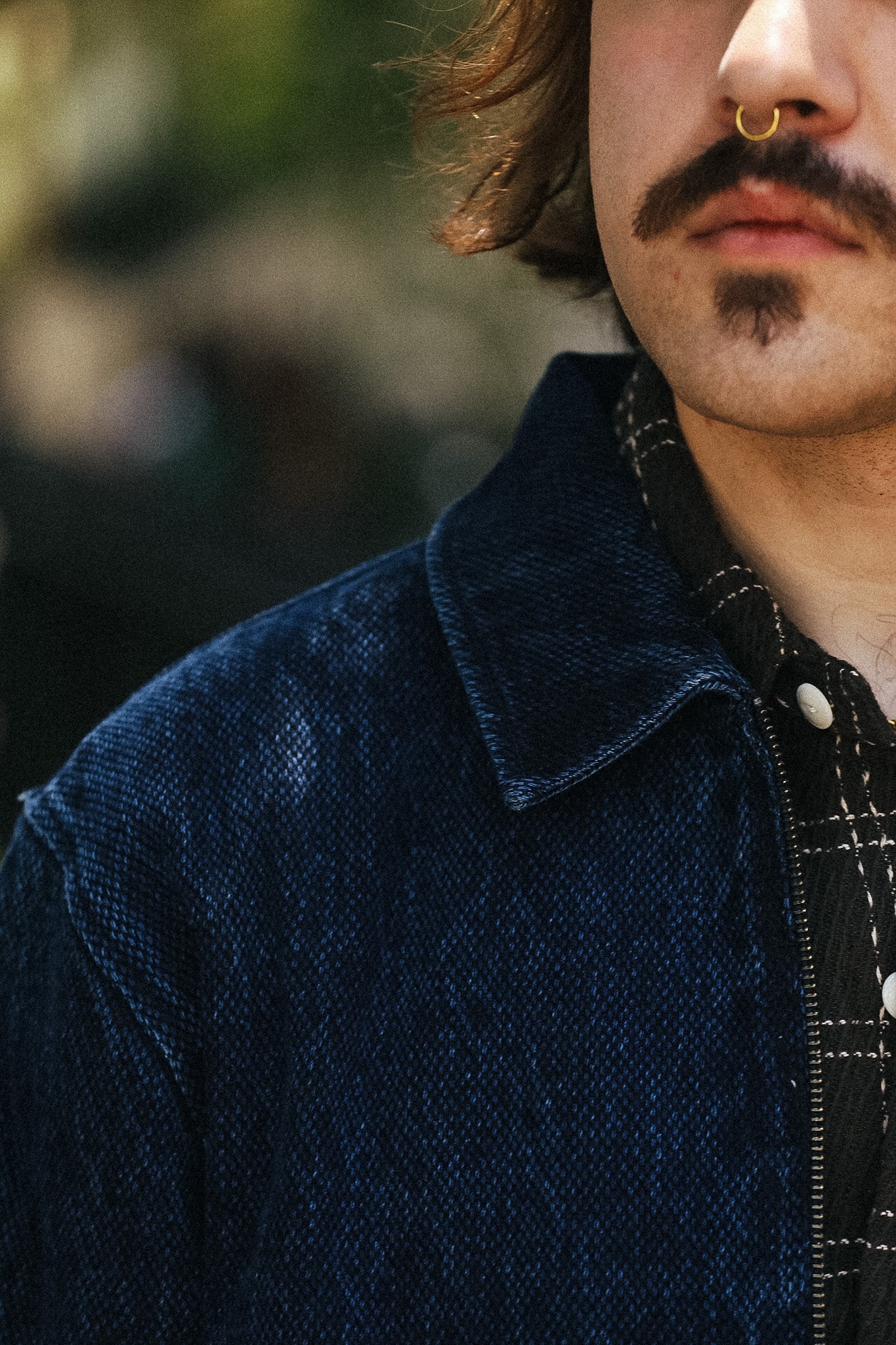 Close-up of a person wearing a dark navy textured overshirt over a black and white plaid shirt, with a gold nose ring and a mustache.
