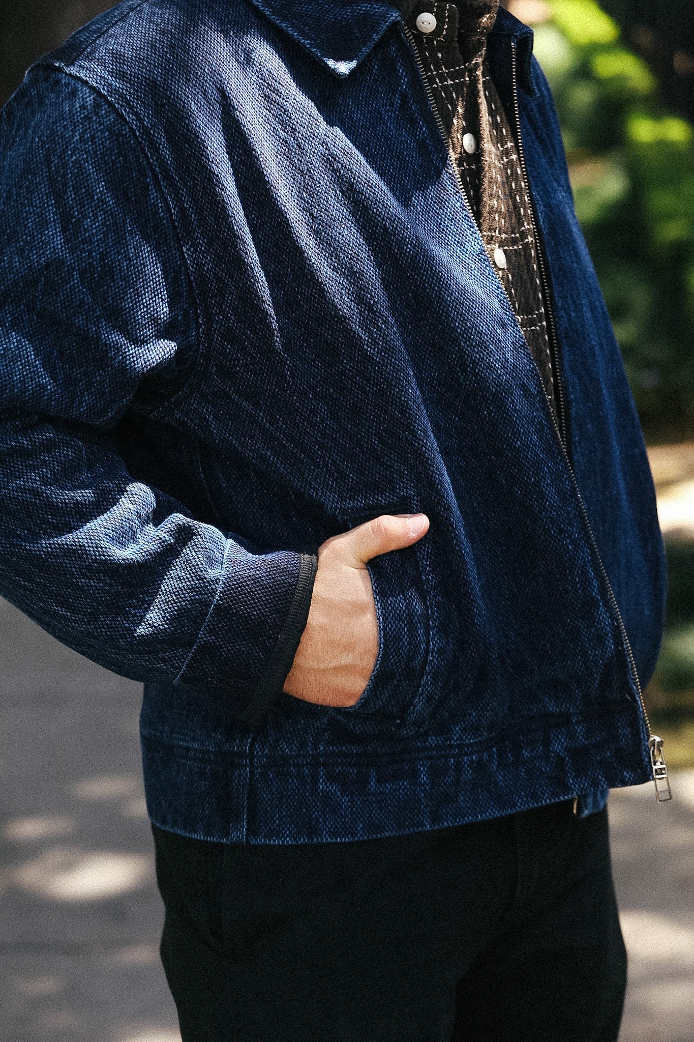 A person wearing a dark indigo denim jacket over a patterned shirt, standing outdoors with one hand in the pocket.