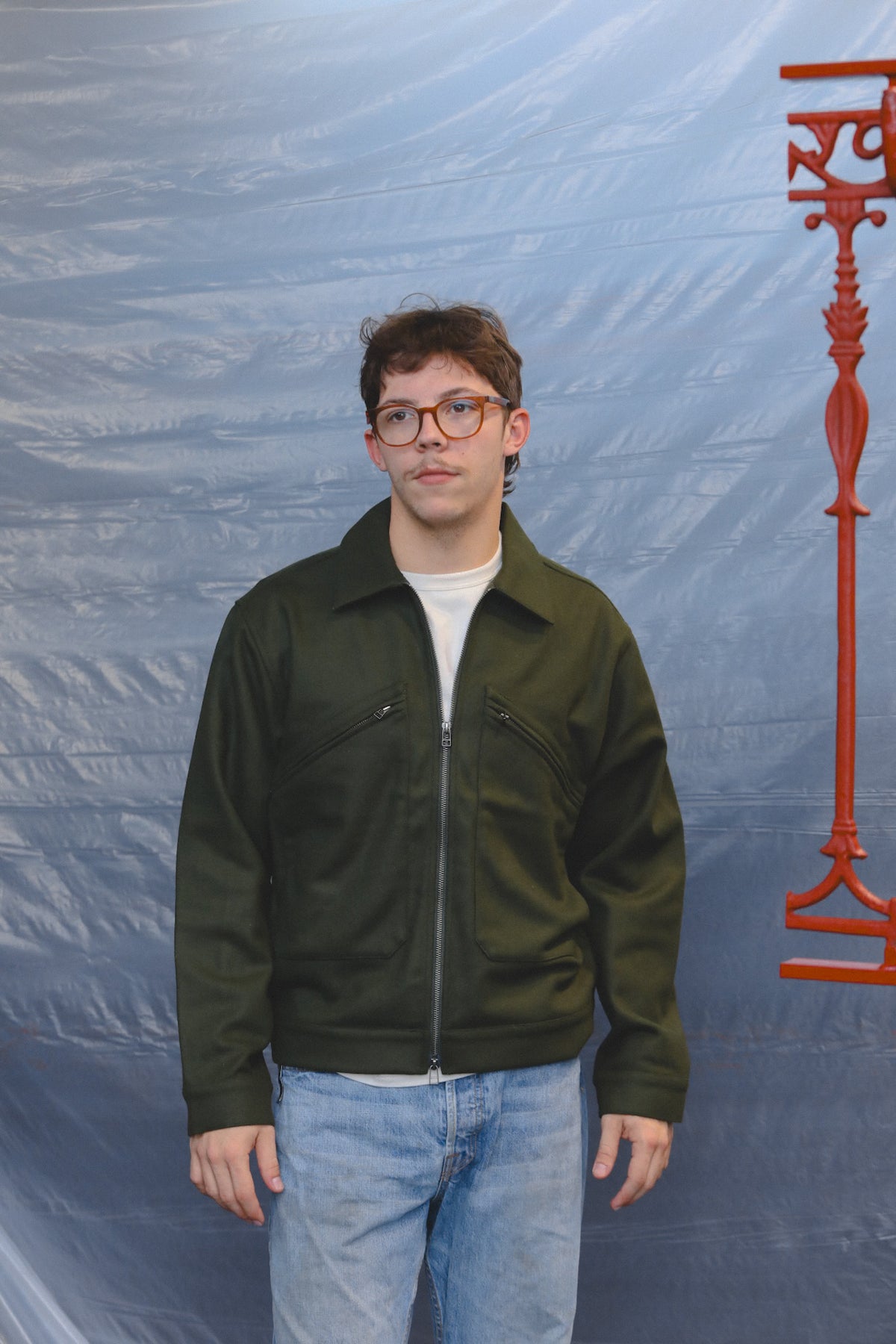 A man wearing a dark olive green zip-up jacket over a white t-shirt and light blue jeans, standing in front of a blue textured backdrop with a red decorative object on the right.