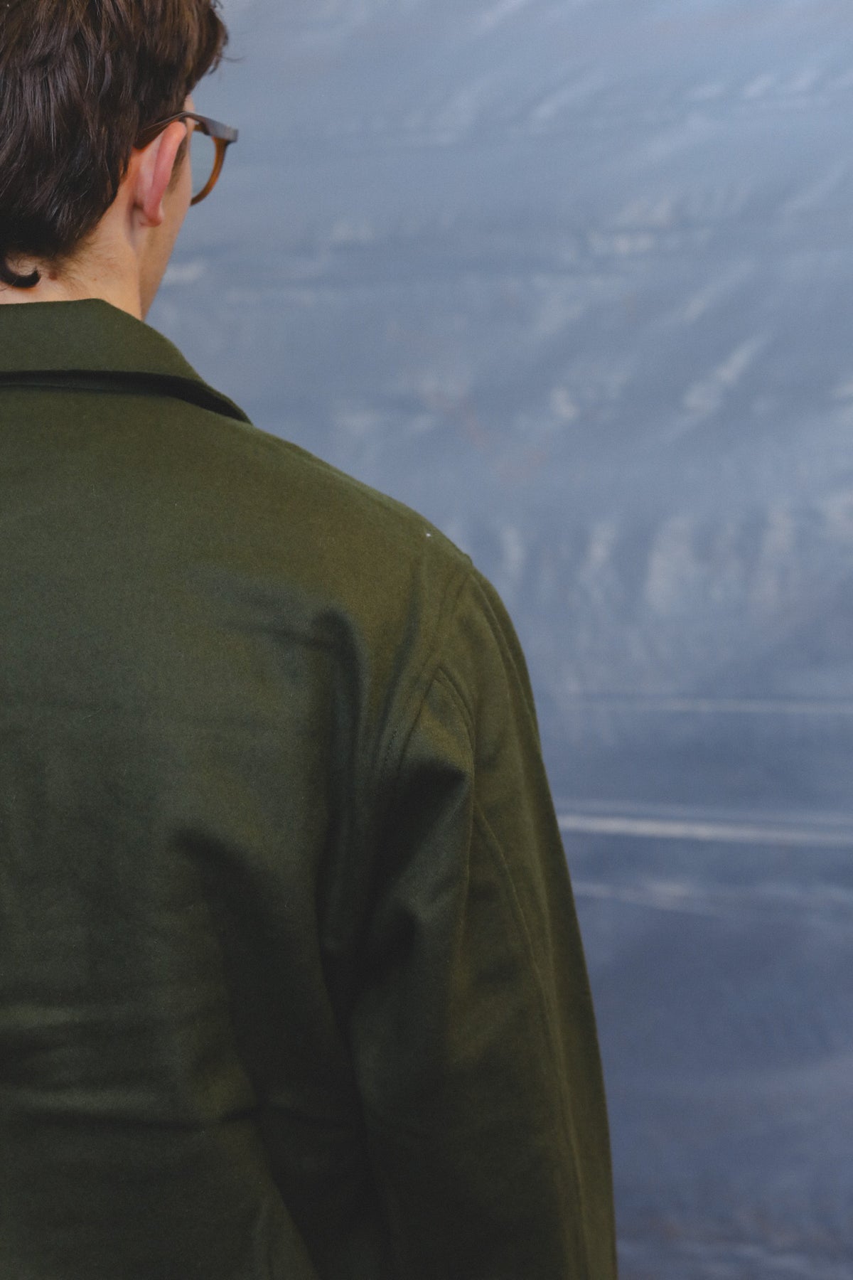A person viewed from behind wearing a dark olive green jacket, looking out toward a blurred blue-gray landscape or water scene.