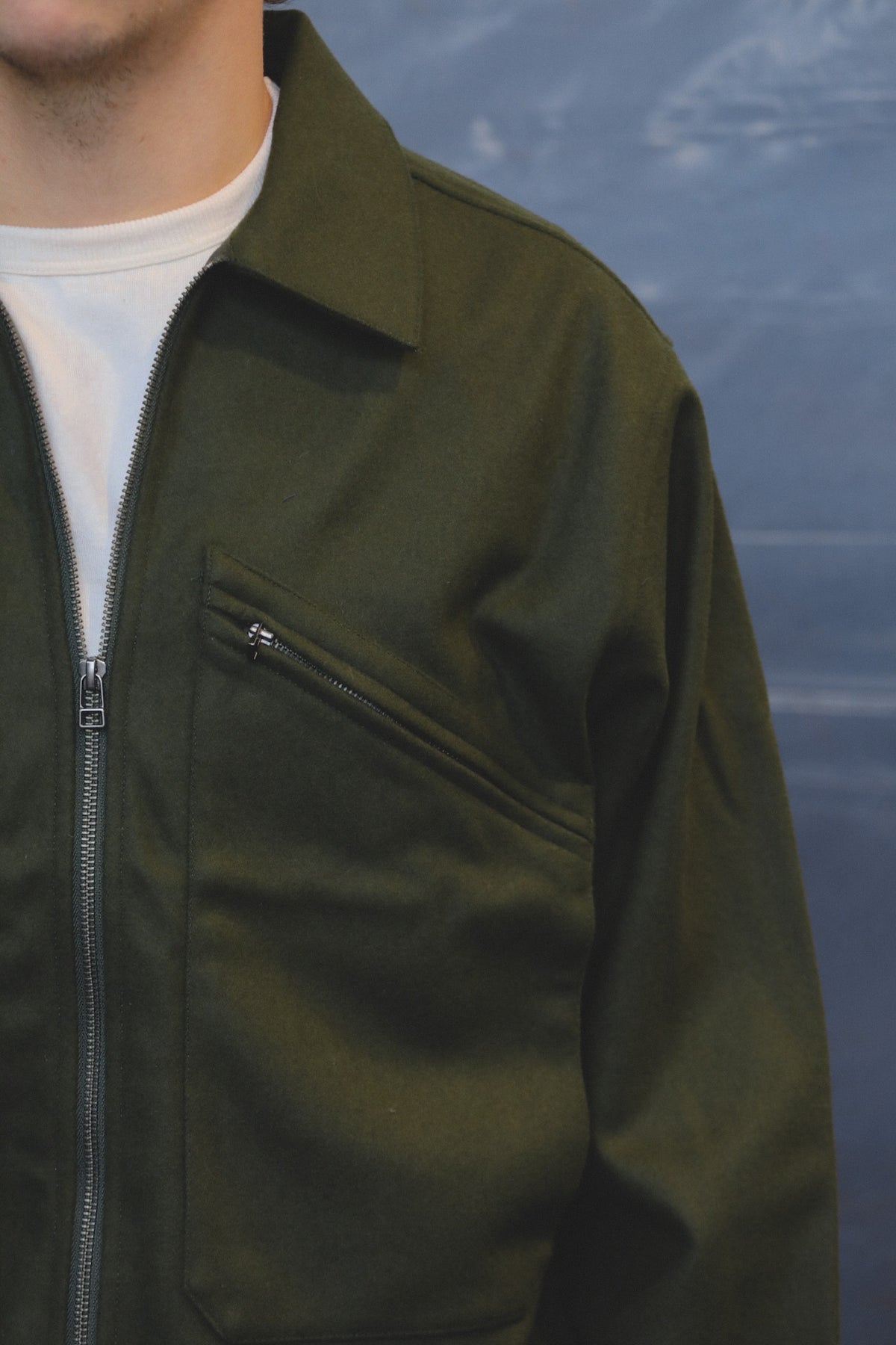 Close-up of a person wearing an olive green zip-front jacket over a white shirt, with a diagonal chest pocket and zipper detail visible.