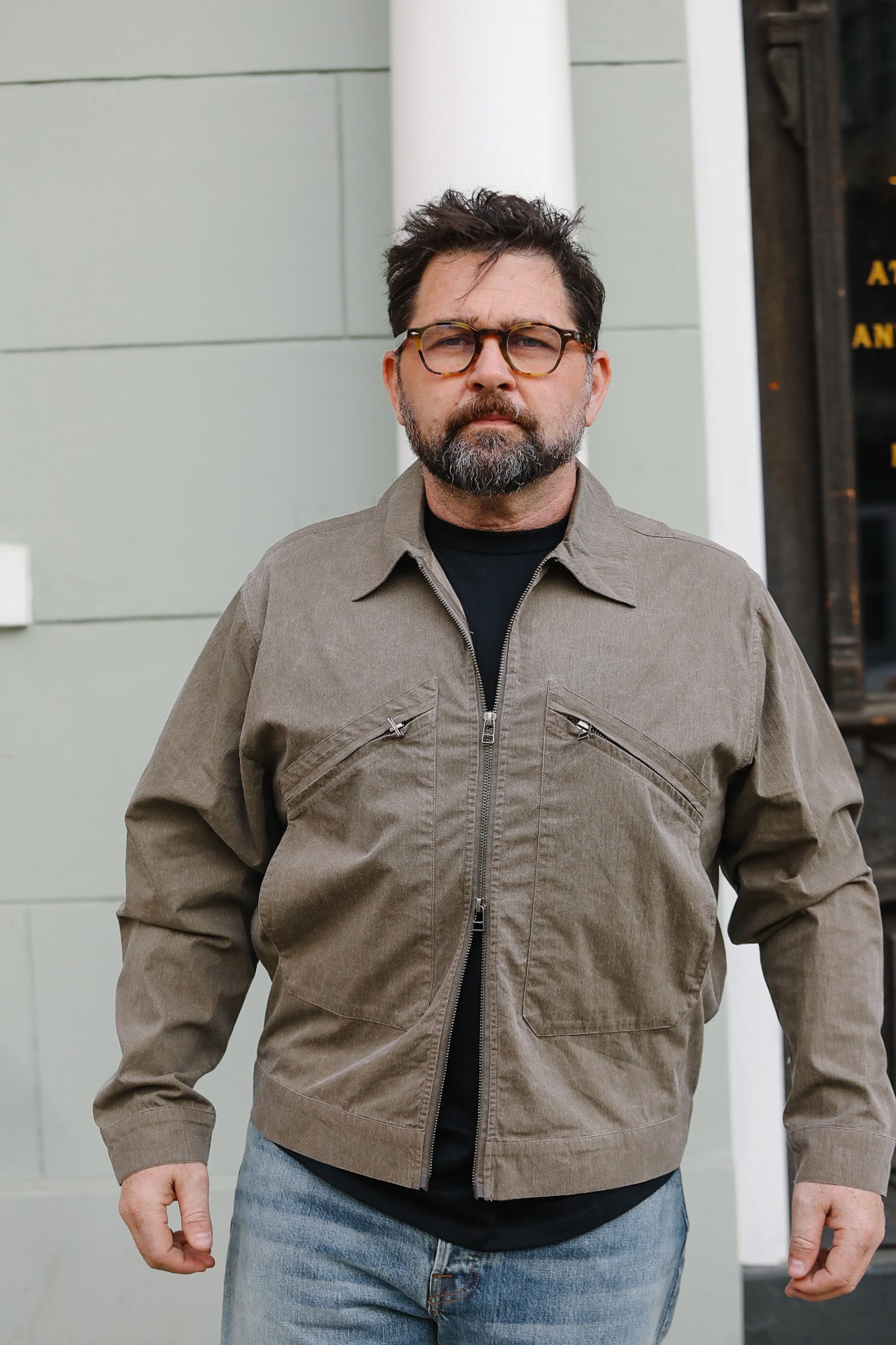 Man wearing a brown jacket and glasses standing against a gray wall.