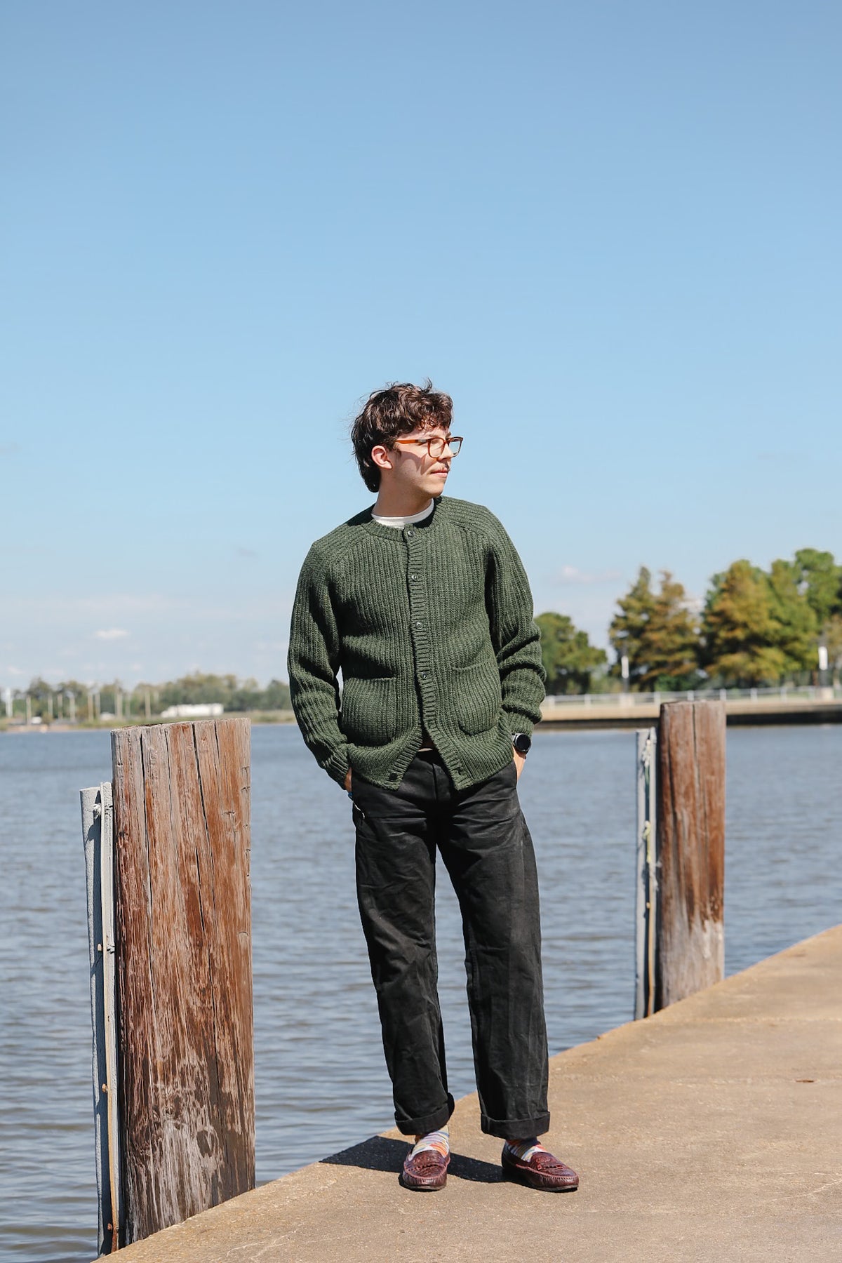 A man wearing a dark green ribbed knit zip-up sweater, black pants, and burgundy loafers stands on a wooden dock next to a body of water under a clear blue sky.
