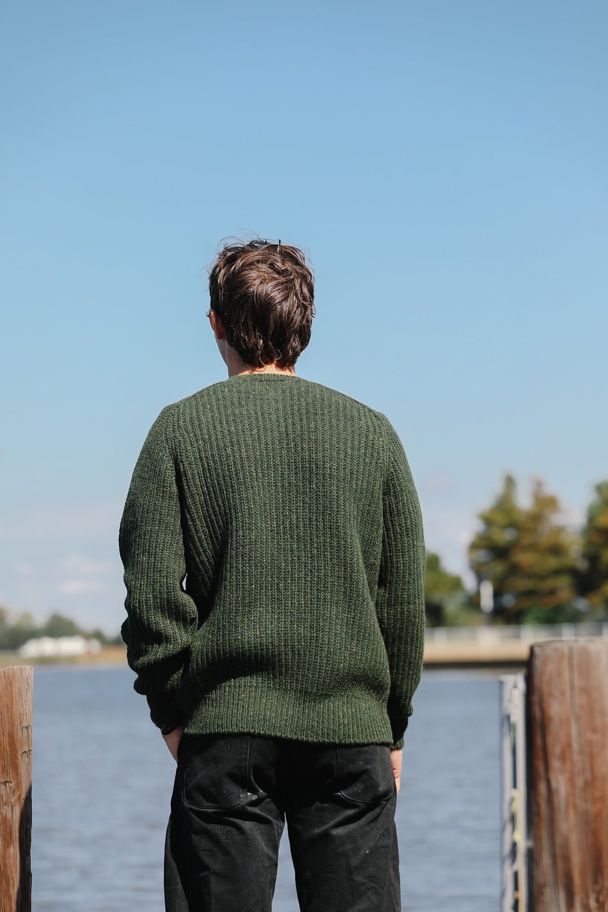 A man viewed from behind wearing a dark green ribbed knit sweater and black pants, standing outdoors by a body of water under a clear blue sky.