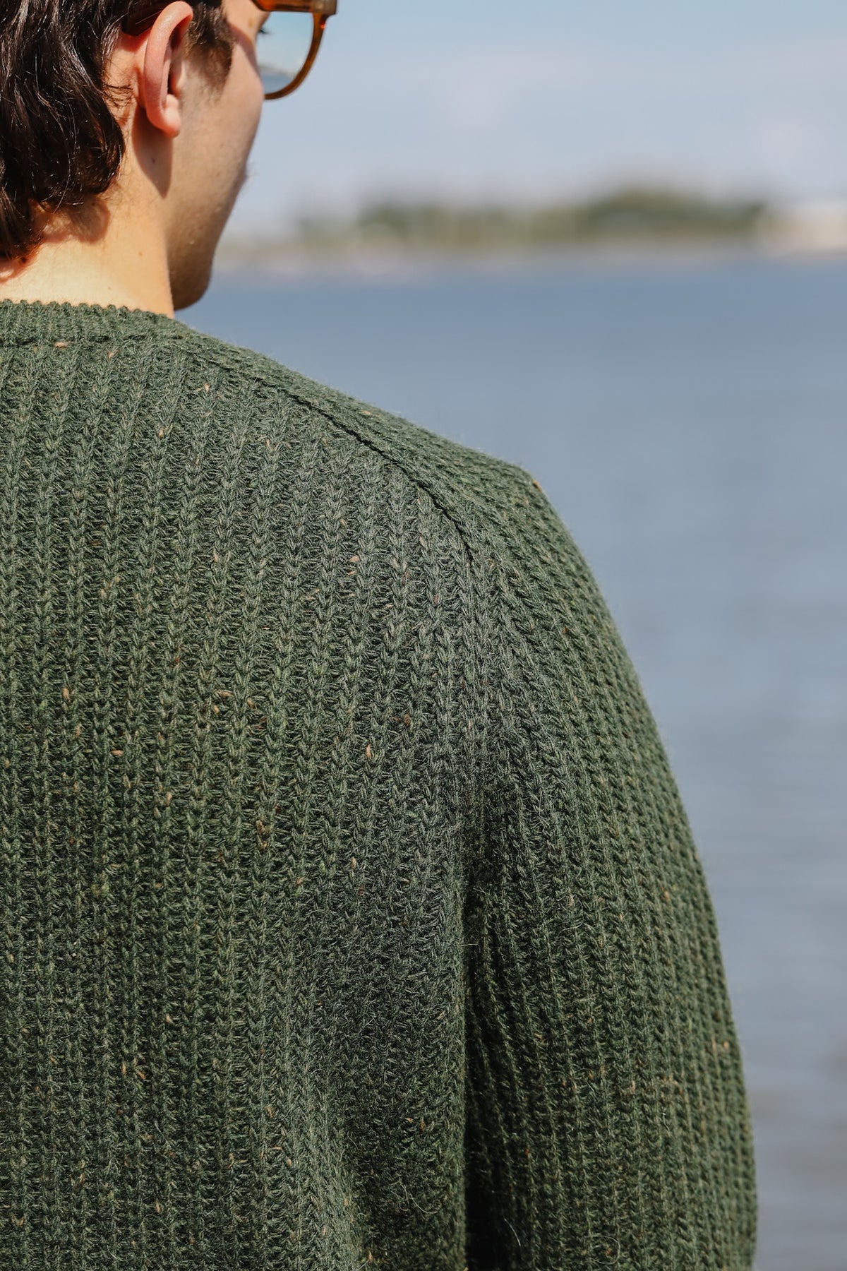 A man wearing a textured olive green knit sweater, shown from behind as he looks out over a body of water with a blurred shoreline in the background.