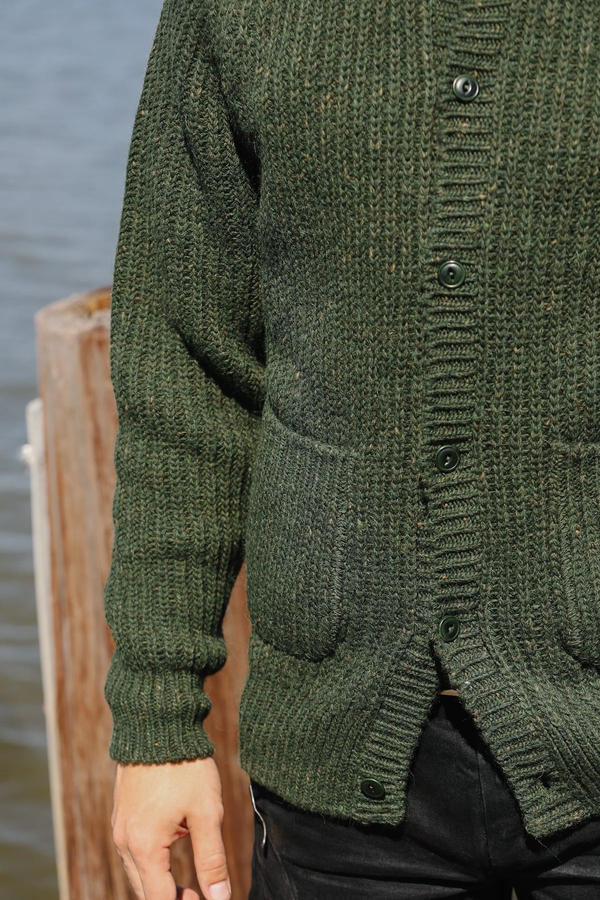 A close-up of a person wearing a dark olive green chunky knit cardigan with dark buttons, standing outdoors near water and a wooden post.