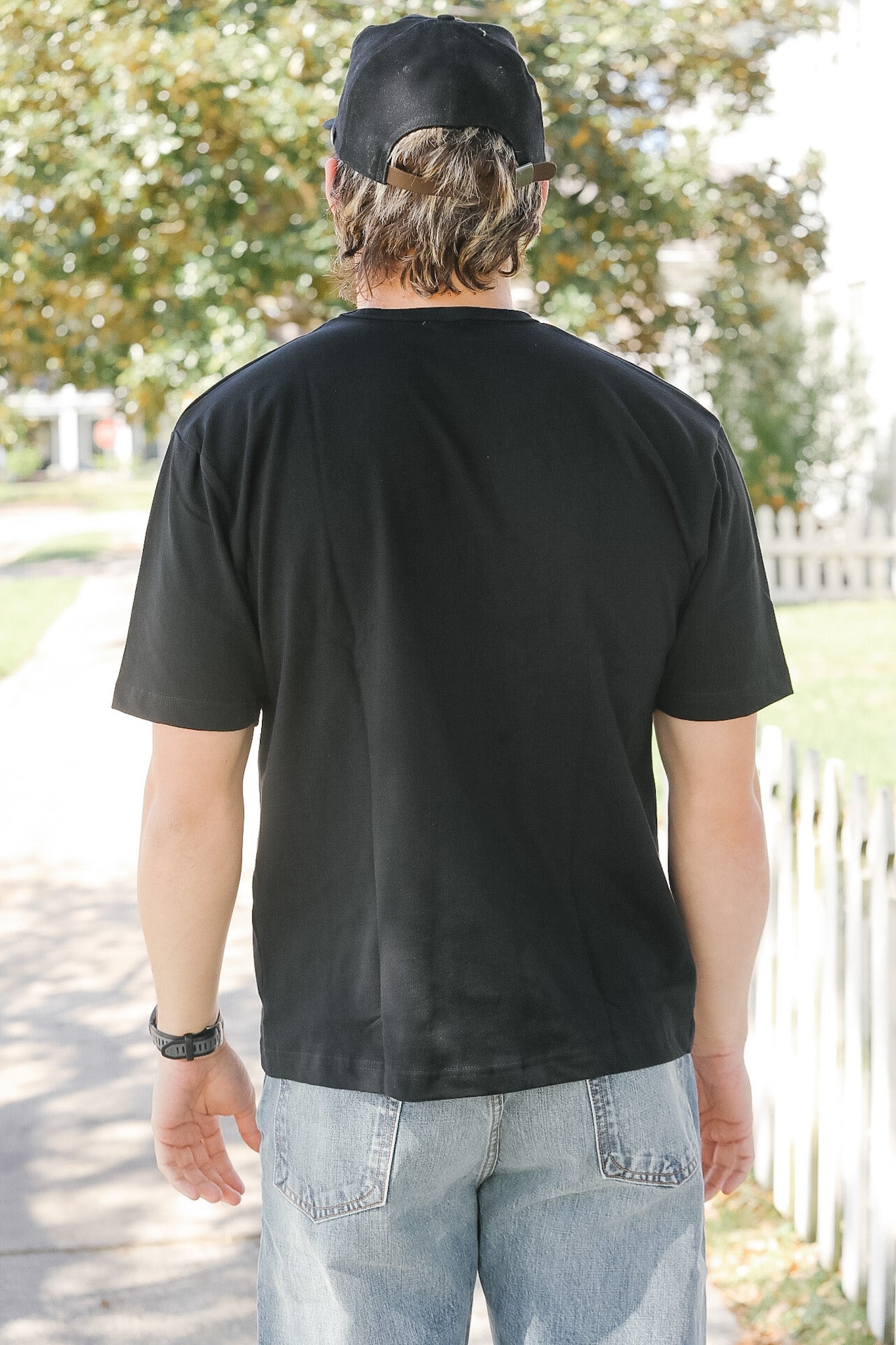 Person wearing a black t-shirt and cap, standing outdoors with trees and a white fence in the background.