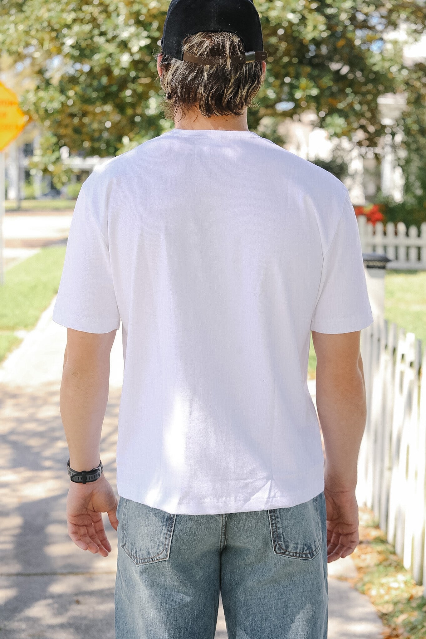 Person wearing a white t-shirt and black cap walking outdoors on a sunny day.
