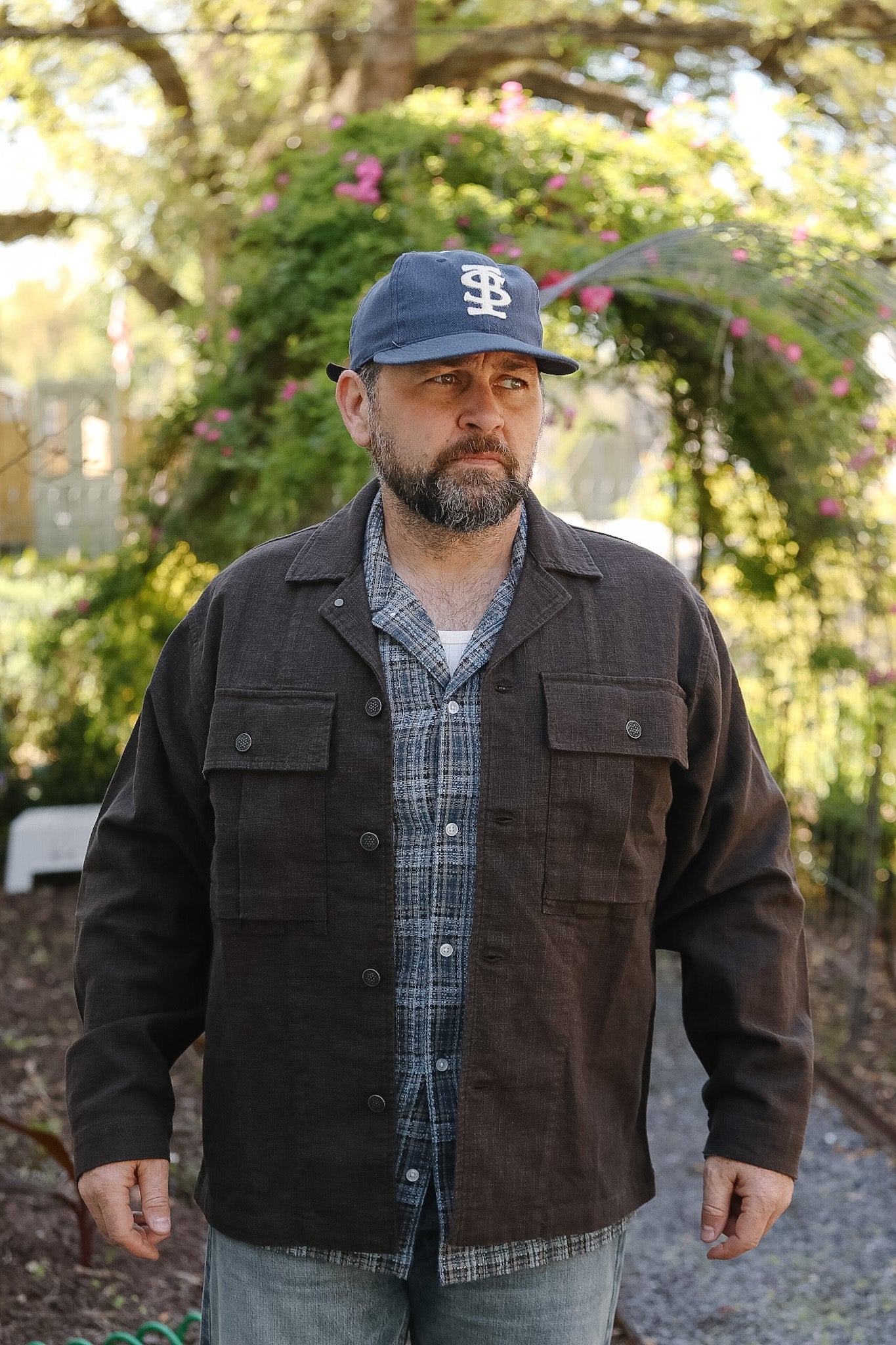 Man wearing a brown jacket, plaid shirt, and blue cap outdoors with greenery in the background