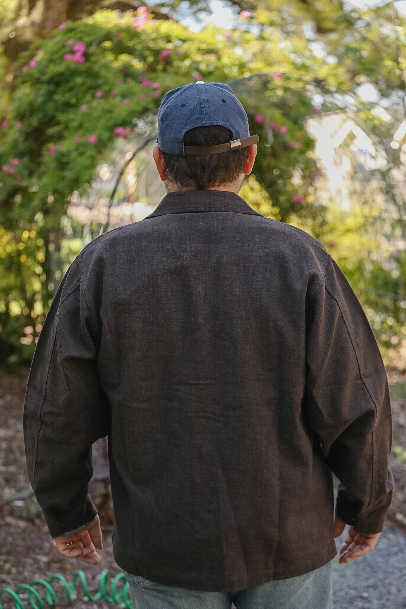 Person wearing a dark jacket and cap walking away from the camera with greenery in the background