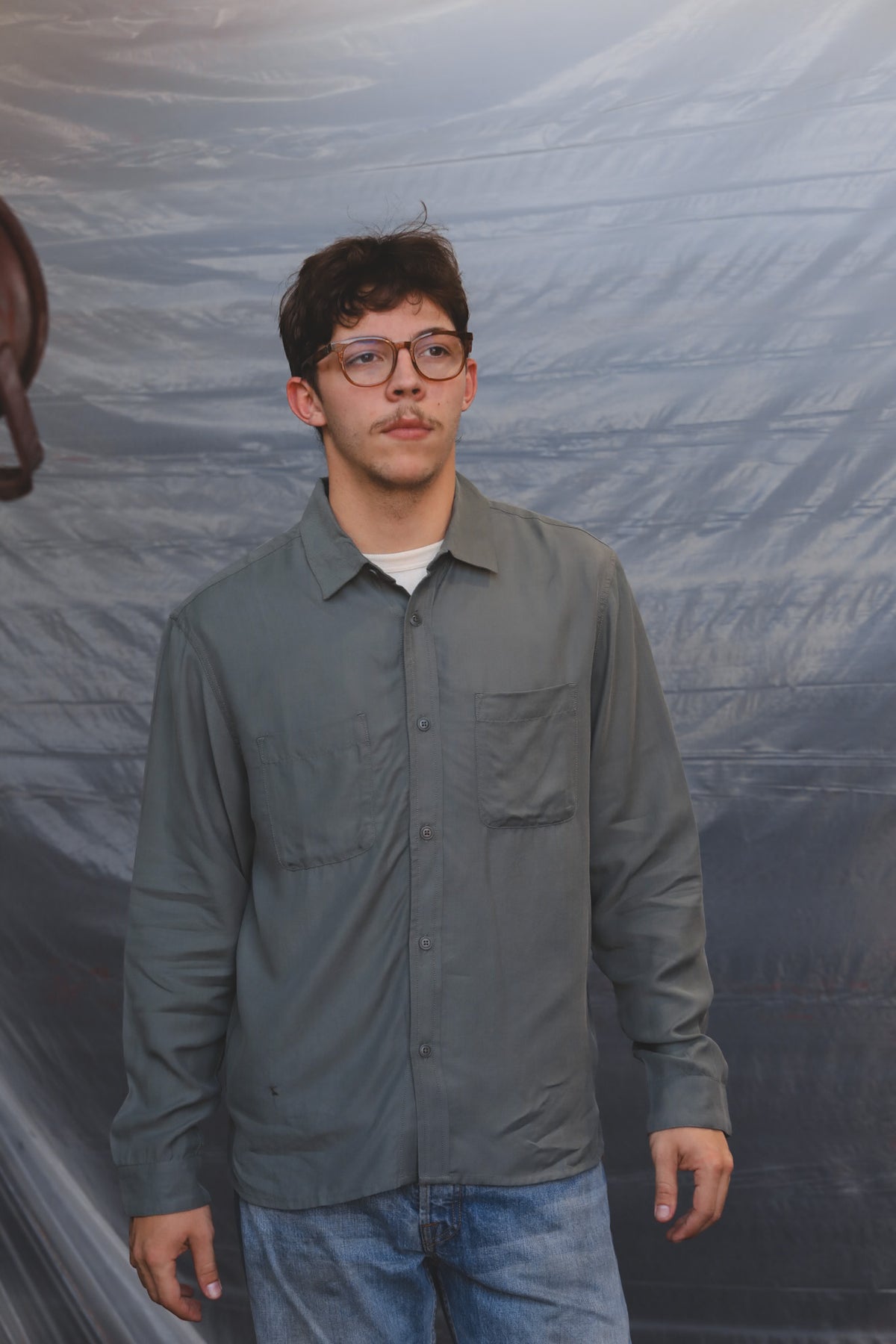 A man wearing a gray long-sleeve button-up shirt and blue jeans, standing in front of a wrinkled gray backdrop.