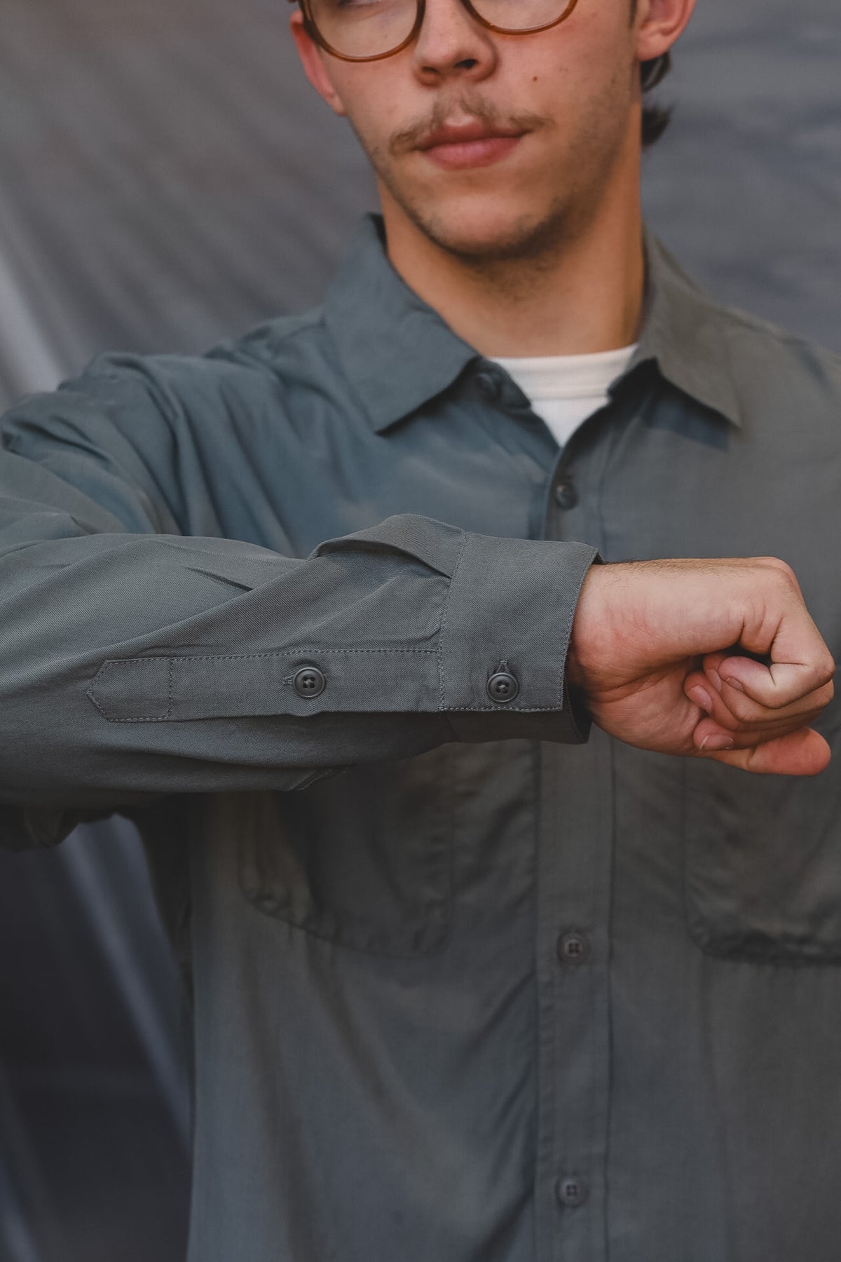A close-up of a man wearing a slate gray button-down shirt over a white undershirt, with his arm raised to show the cuff detailing.