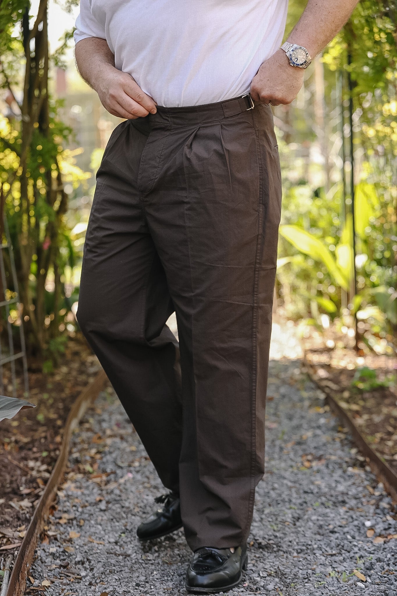 Person wearing brown pants and a white shirt standing on a gravel path with greenery in the background