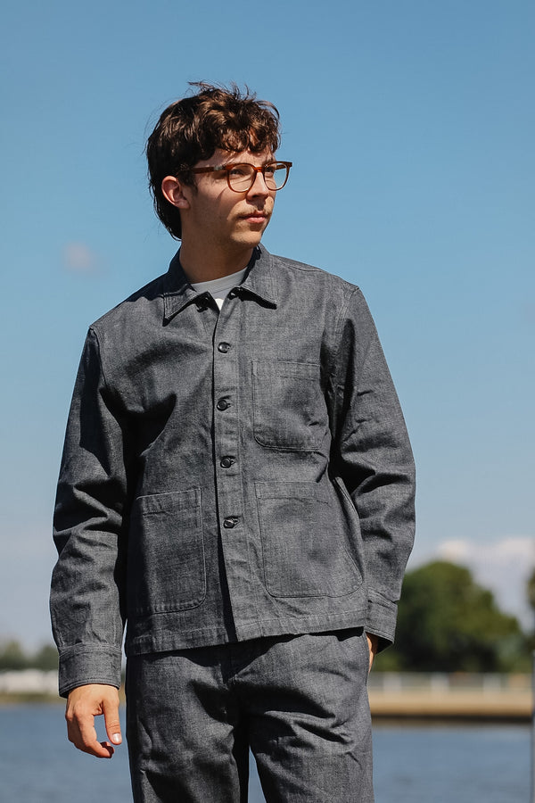 A man wearing a dark gray linen work jacket and matching trousers, standing outdoors against a blue sky and water background.